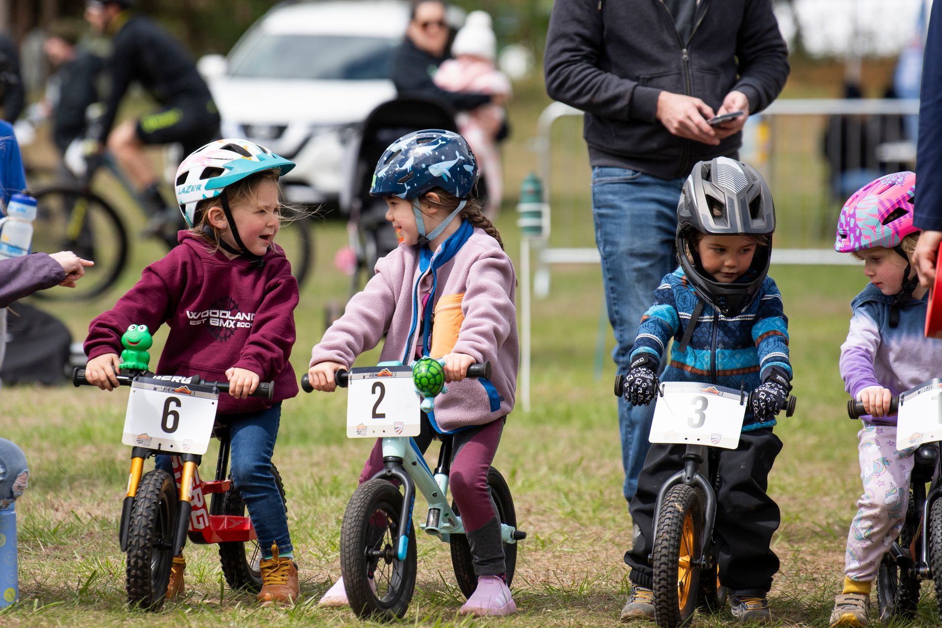 Children on balance bikes at a race, wearing helmets. Numbers on bikes, spectators in background.