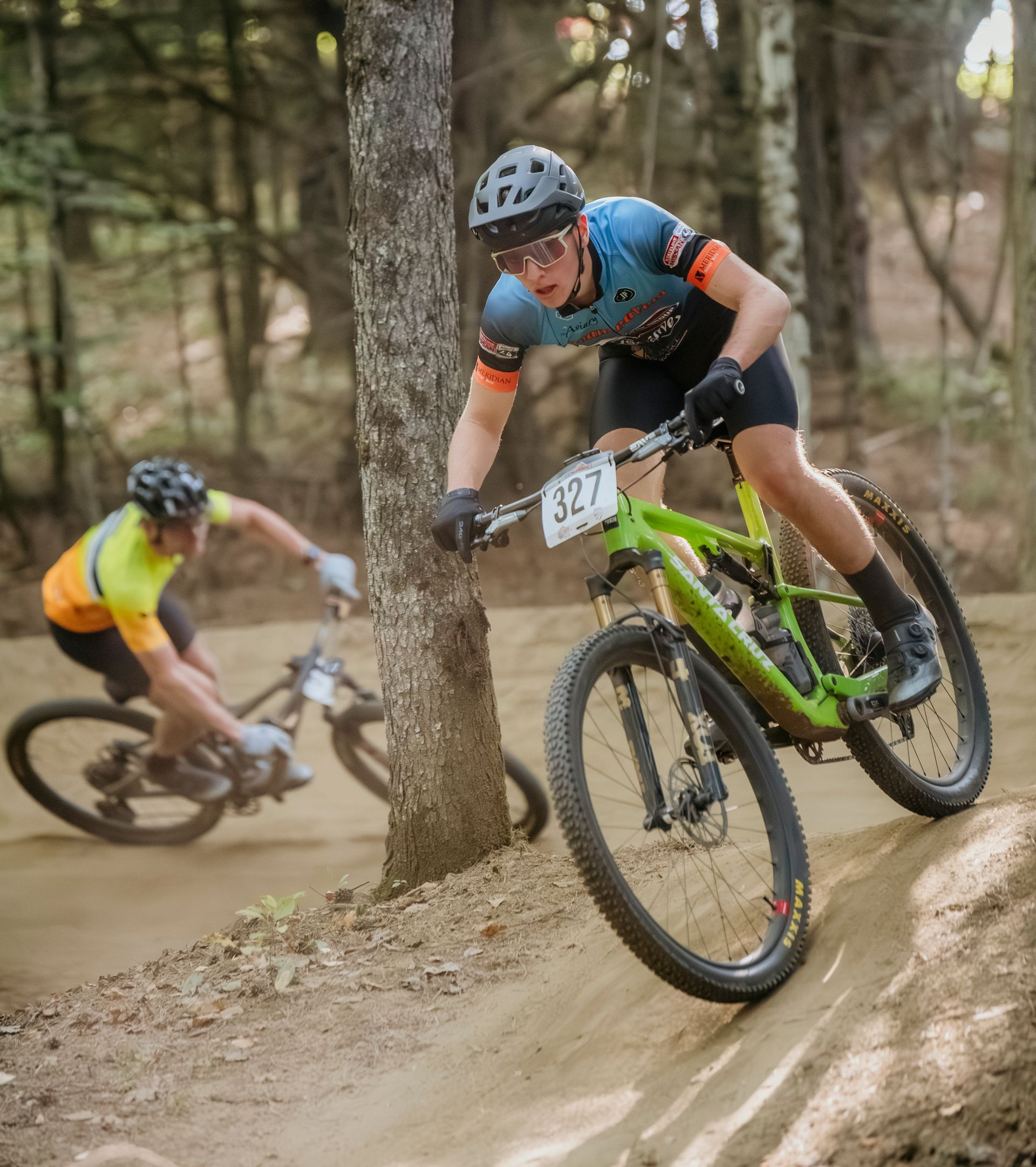 Two mountain bikers racing on a dirt track in a forest. One rider in a blue jersey, leaning, near a tree. Other rider in a yellow jersey, blurred.