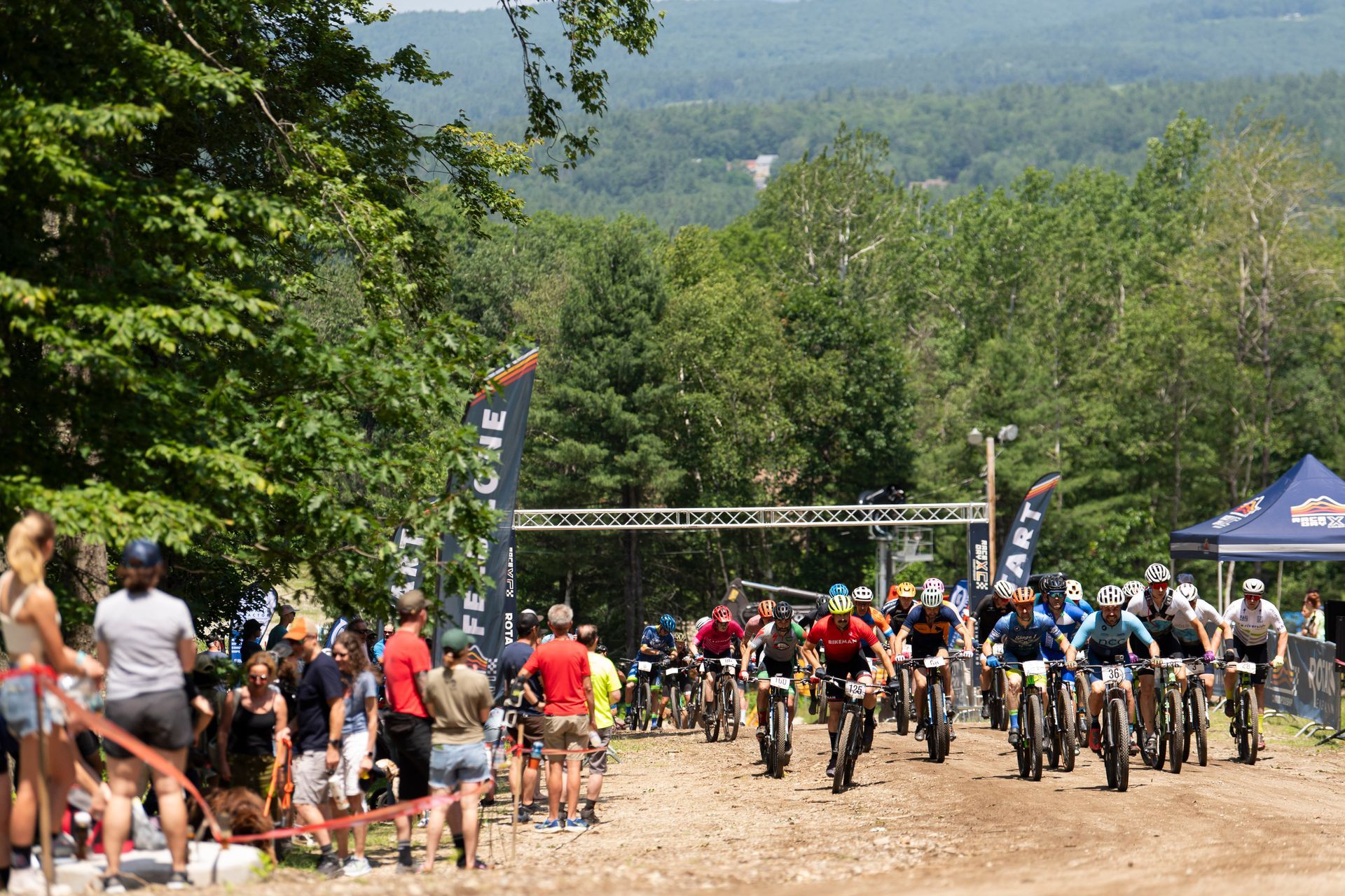 Mountain bikers racing on a dirt path under a metal archway, surrounded by trees and spectators.