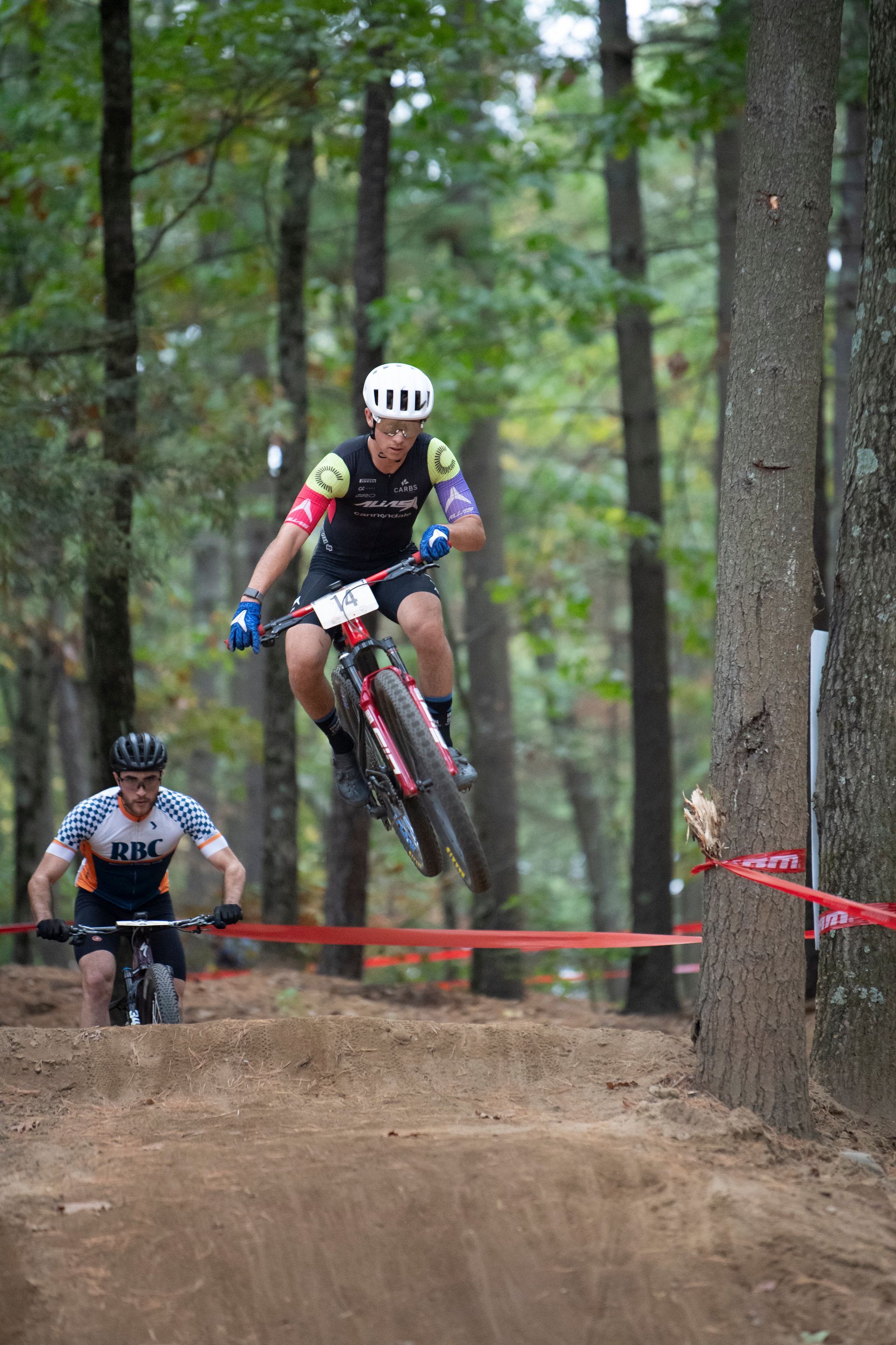 Mountain biker in mid-air over a dirt jump in a forest, another biker watches.