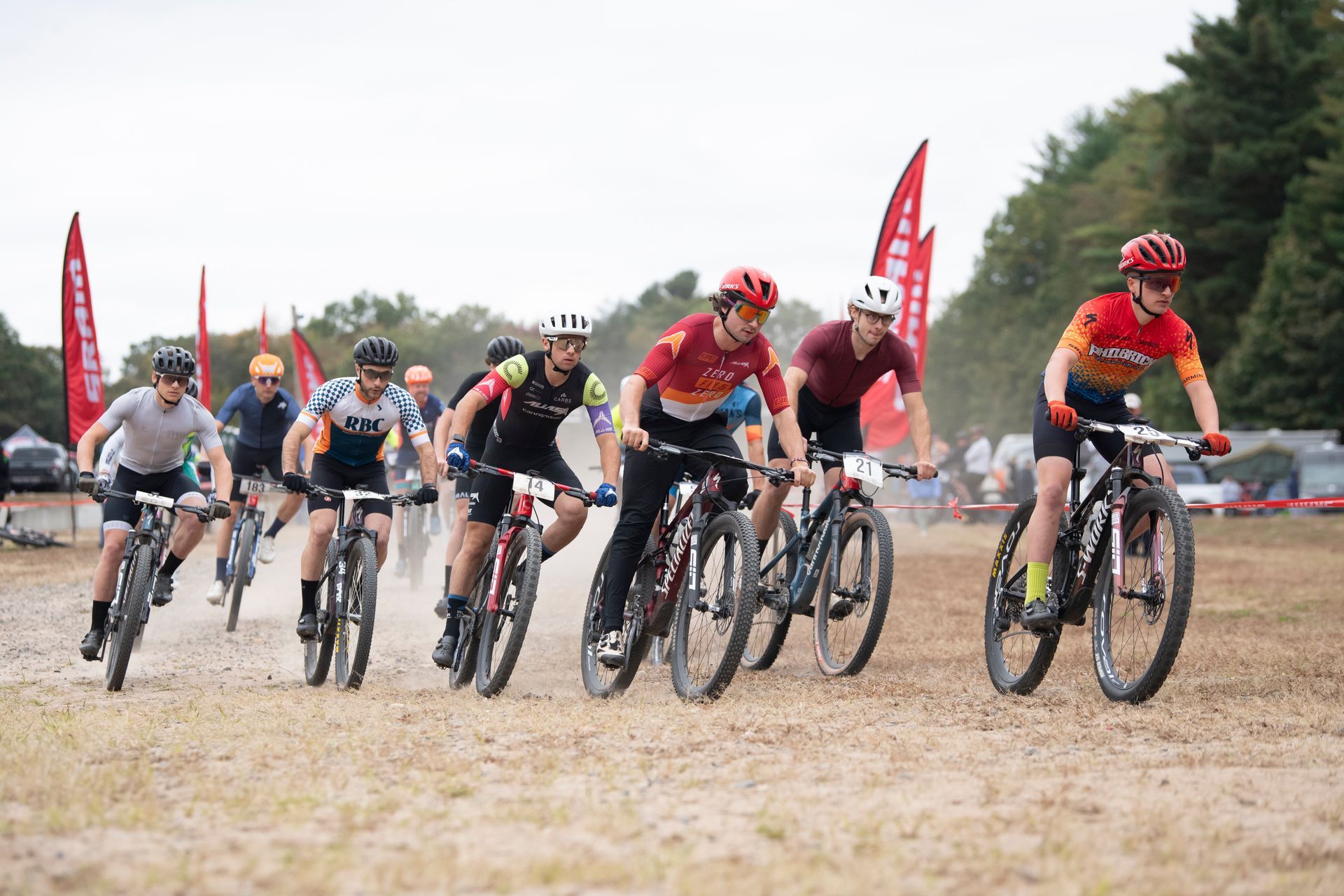 Mountain bike race on a dusty course, riders in helmets and jerseys, red flags in background.