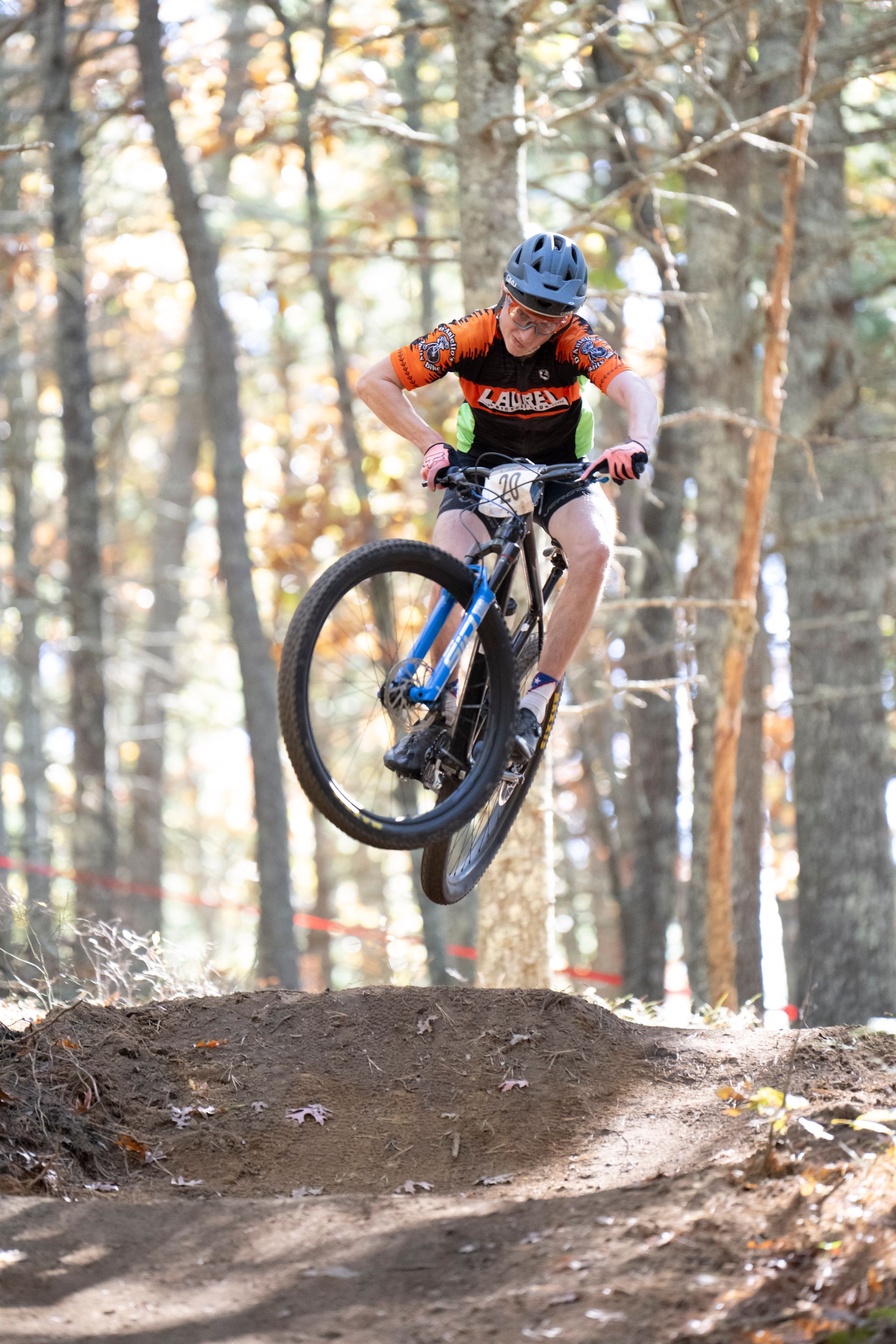 Mountain biker in mid-air over a dirt jump, wearing orange jersey and helmet, in a wooded area.