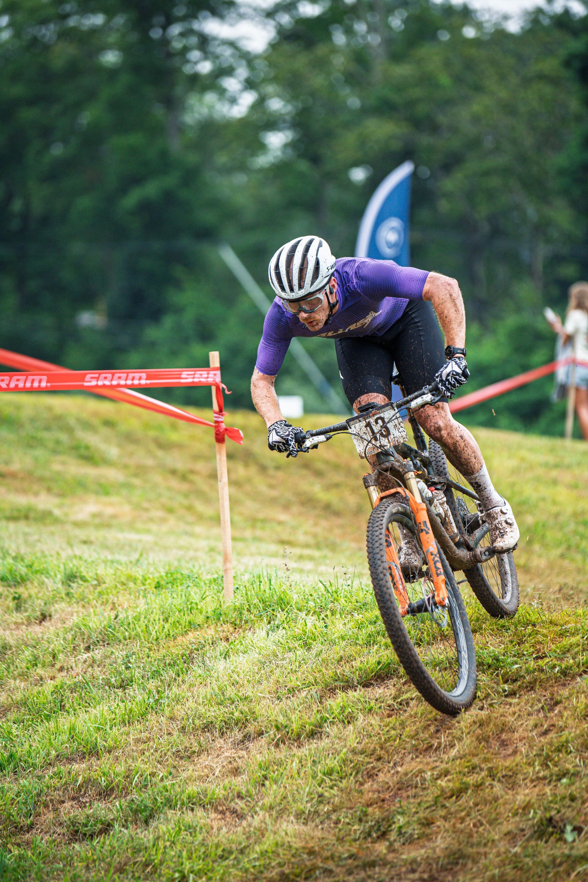 Mountain biker in purple jersey, helmet, descending grassy slope, dirt flying.