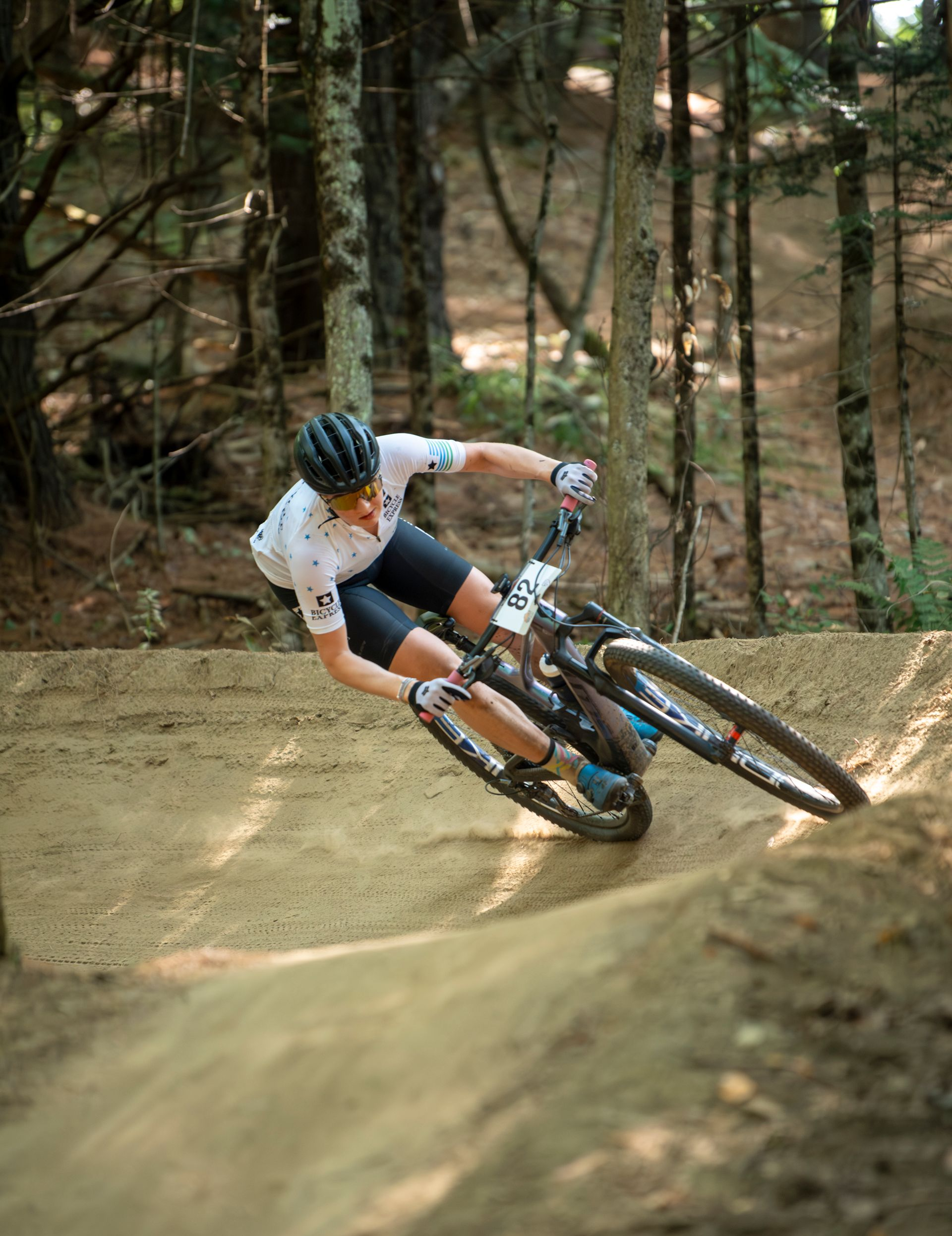 Mountain biker leans into a berm turn on a dirt trail, trees in the background.