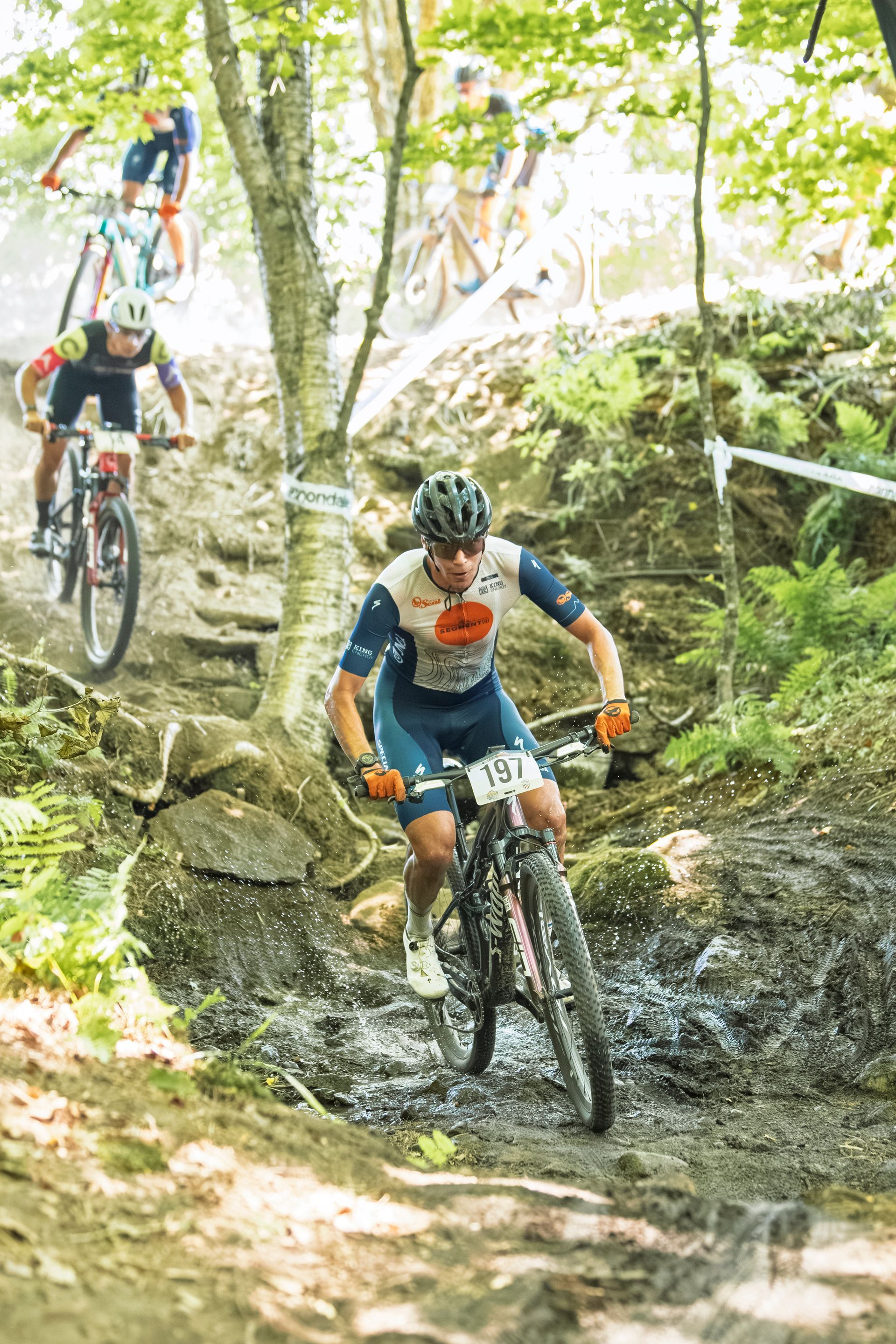Mountain biker navigates a rocky, muddy trail, wearing blue and orange uniform, other riders visible.