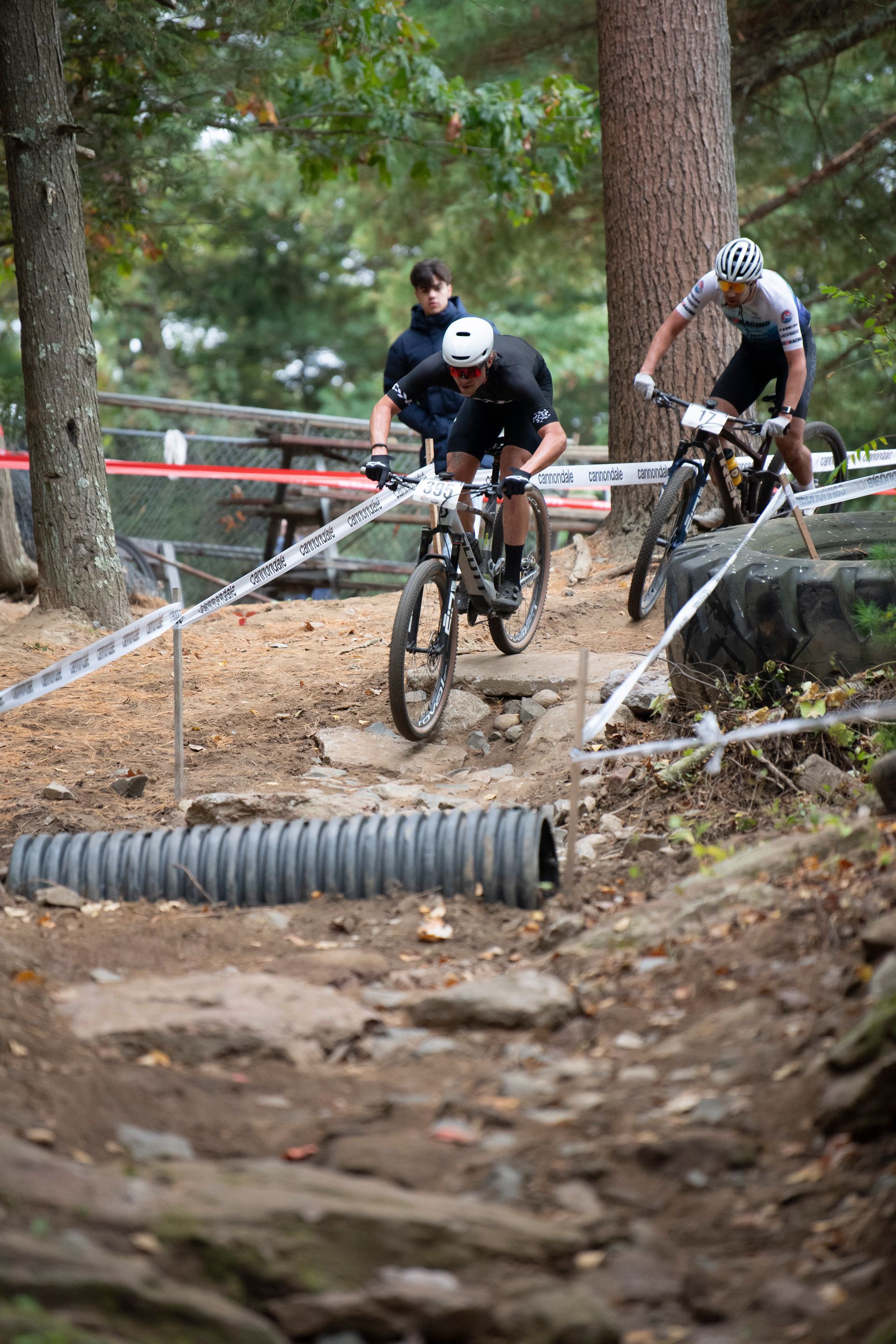 Mountain bikers navigating a rocky trail, one in the foreground, another behind, near a wooded area.