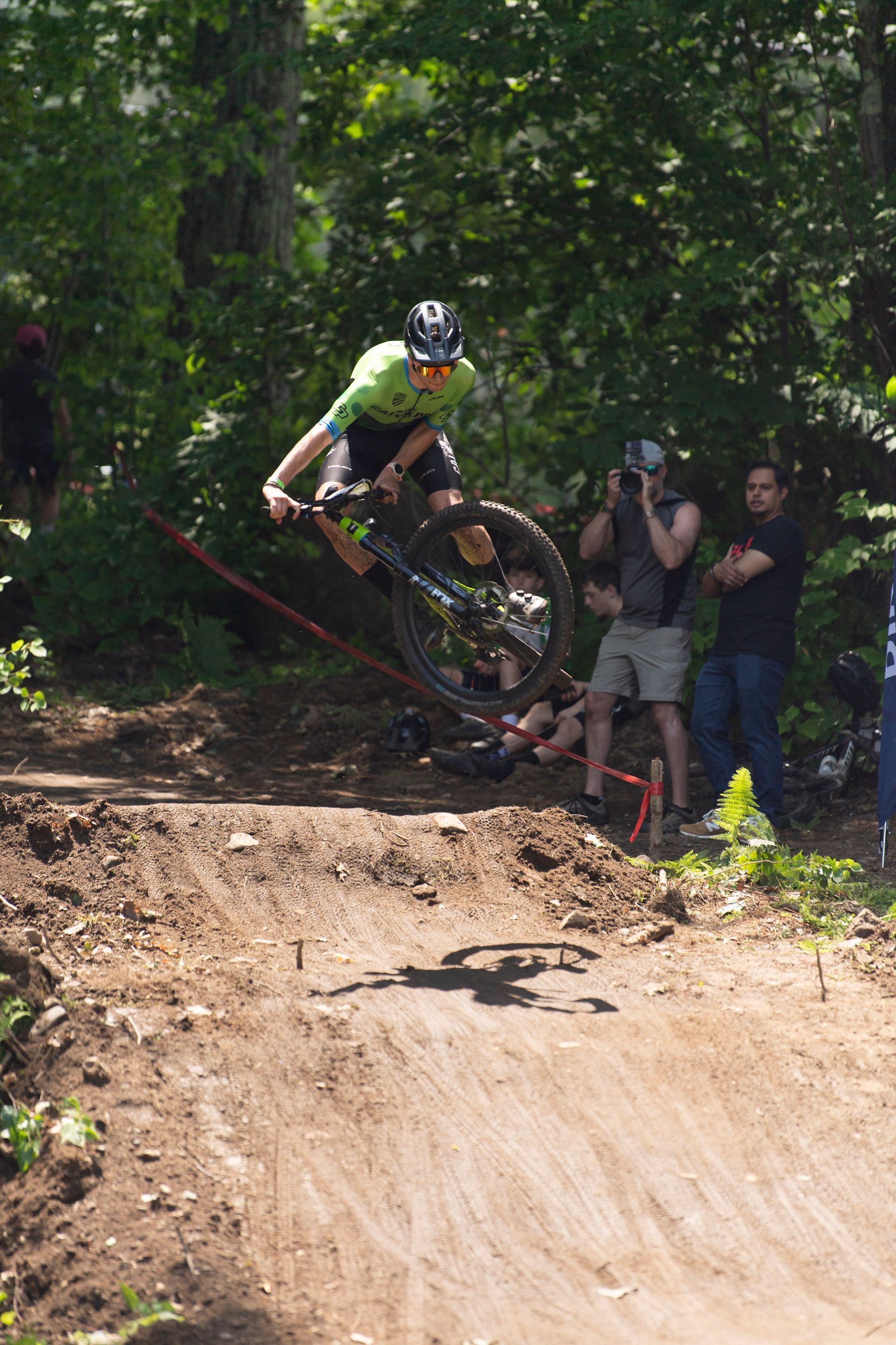 Mountain biker in green gear jumps dirt ramp; two men watch in sunny forest.