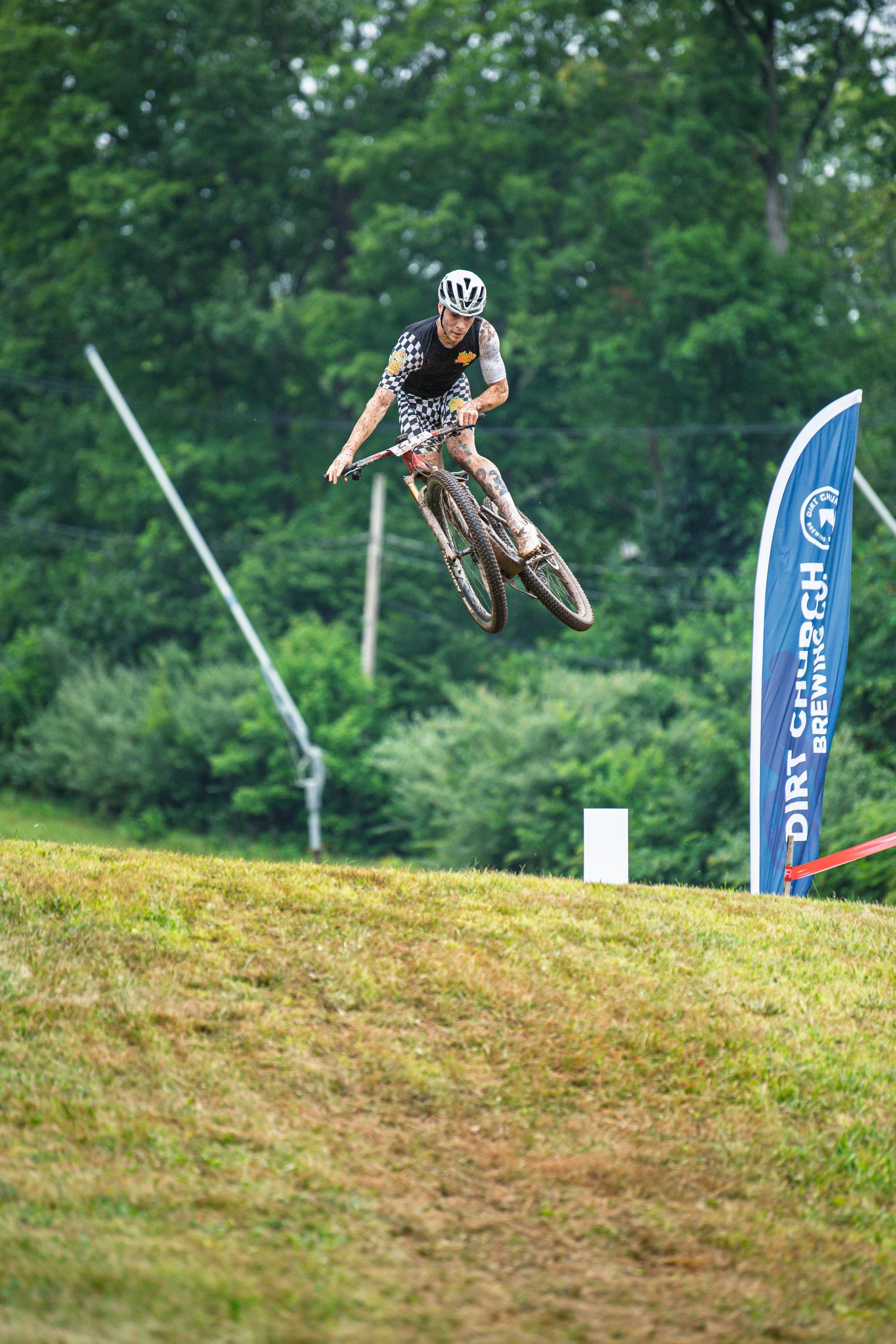 Mountain biker airborne on a dirt jump, grey sky, green grass, trees in the background.