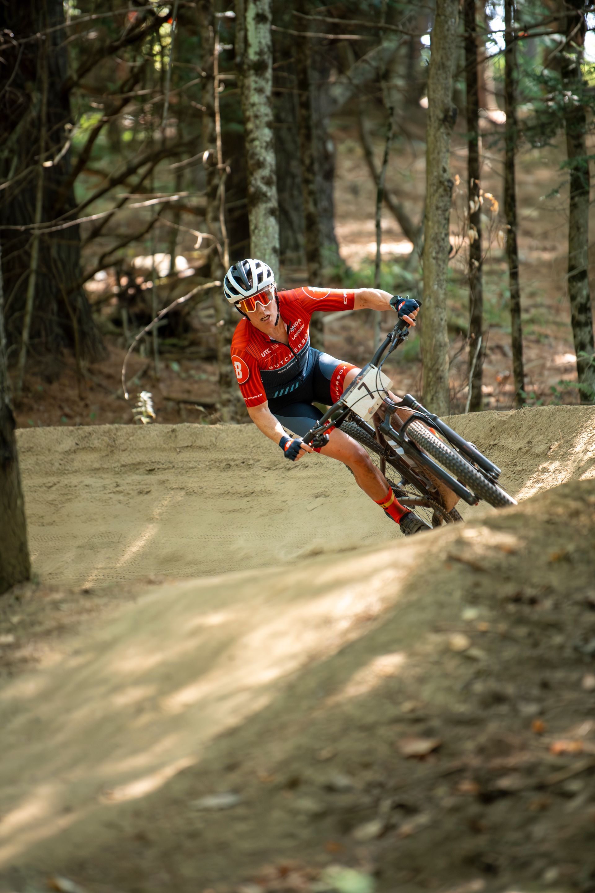 Mountain biker in red and blue gear, leaning into a turn on a dirt trail in a forest.