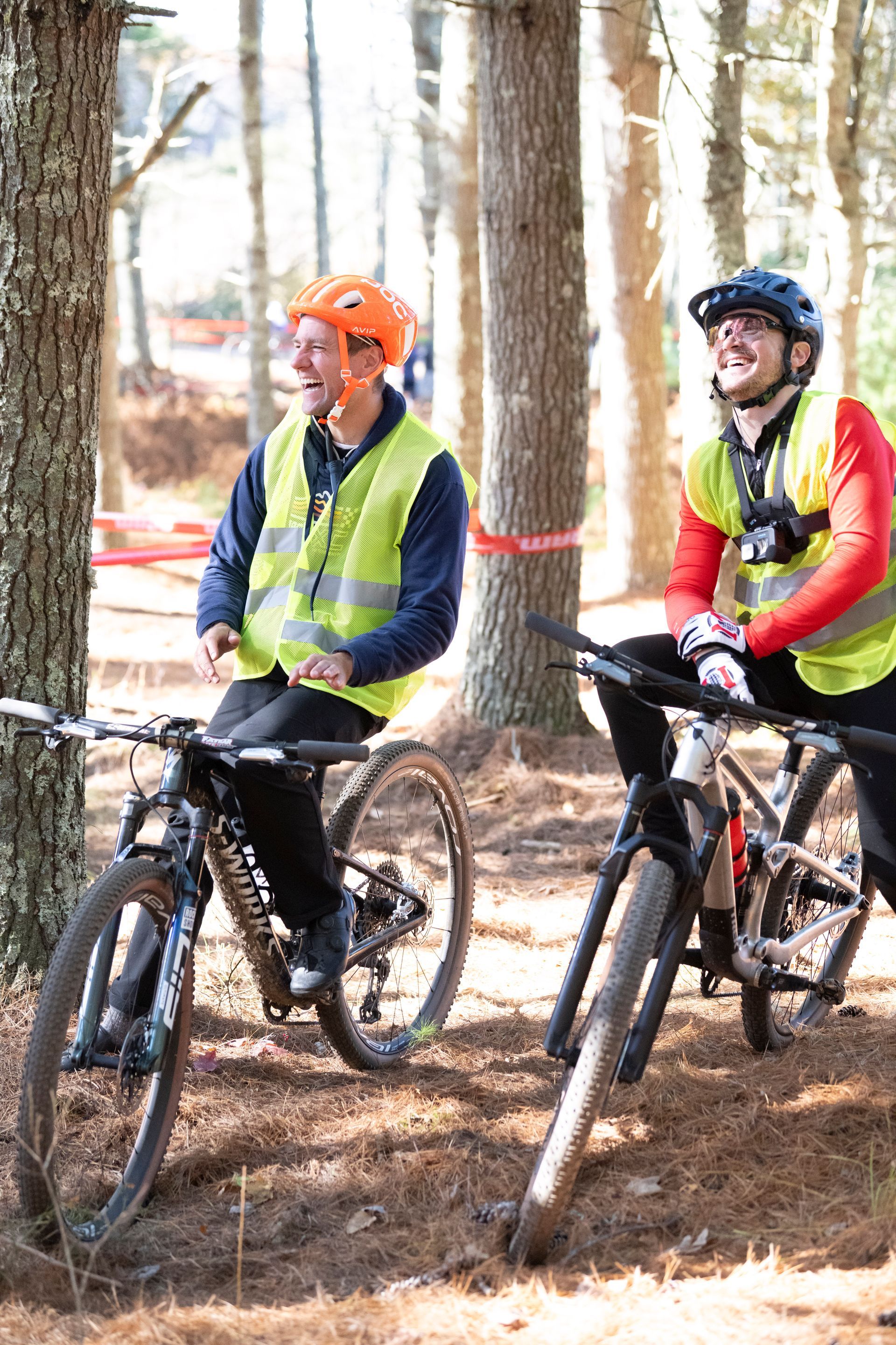 Two cyclists in reflective vests and helmets laugh in a wooded area, bikes at the ready.