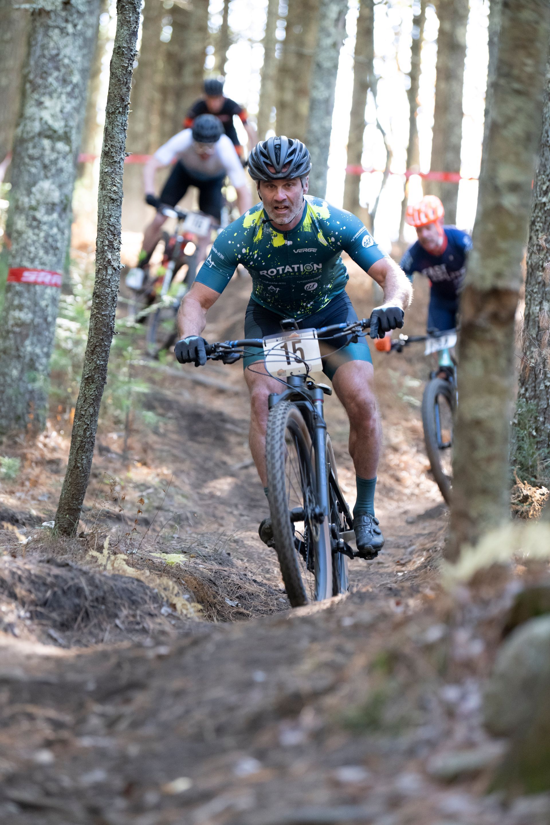 Cyclist on mountain bike navigating a wooded trail, other racers follow behind.