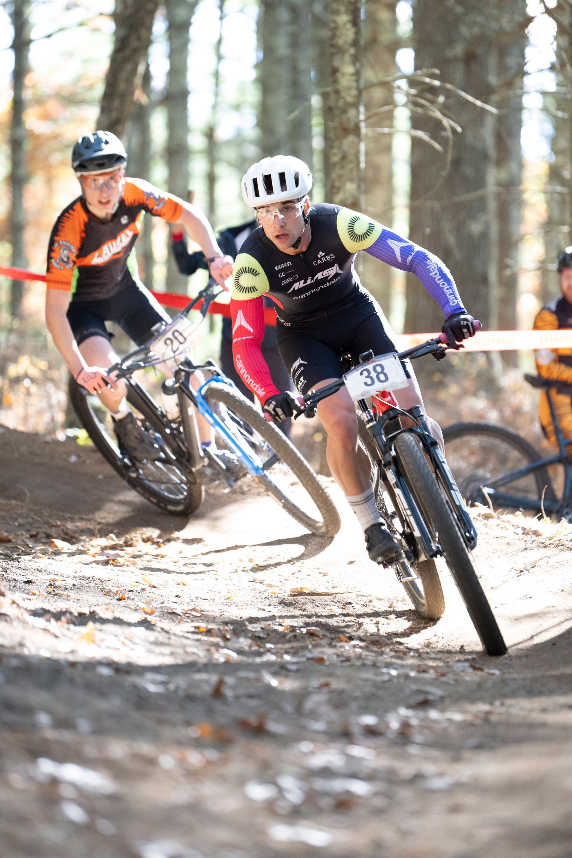 Two cyclists racing on a dirt path in a wooded area. One in front wears a helmet and black and purple kit.