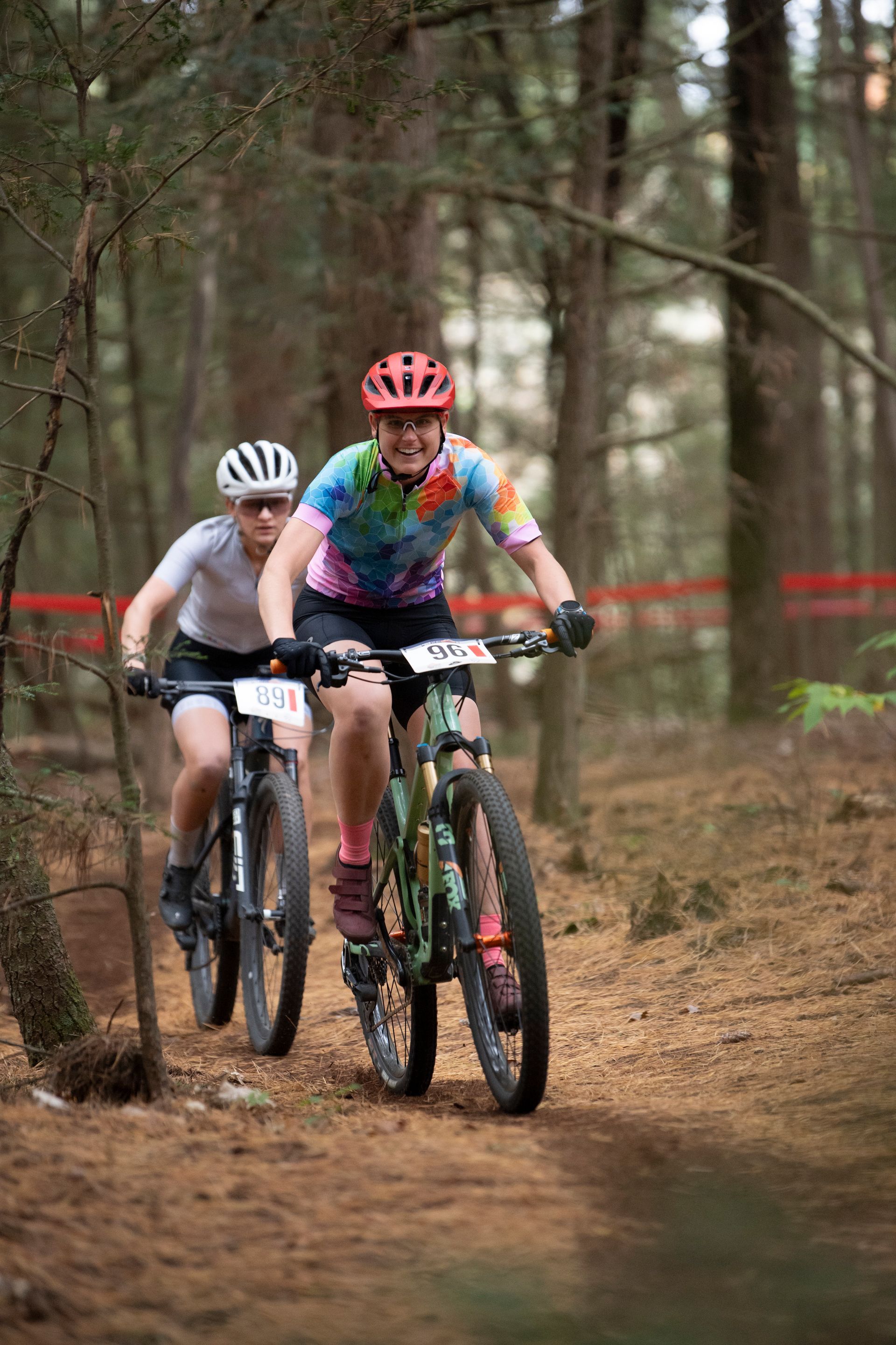 Two cyclists on mountain bikes racing on a dirt trail in a forest. One wears a tie-dye shirt and pink helmet.