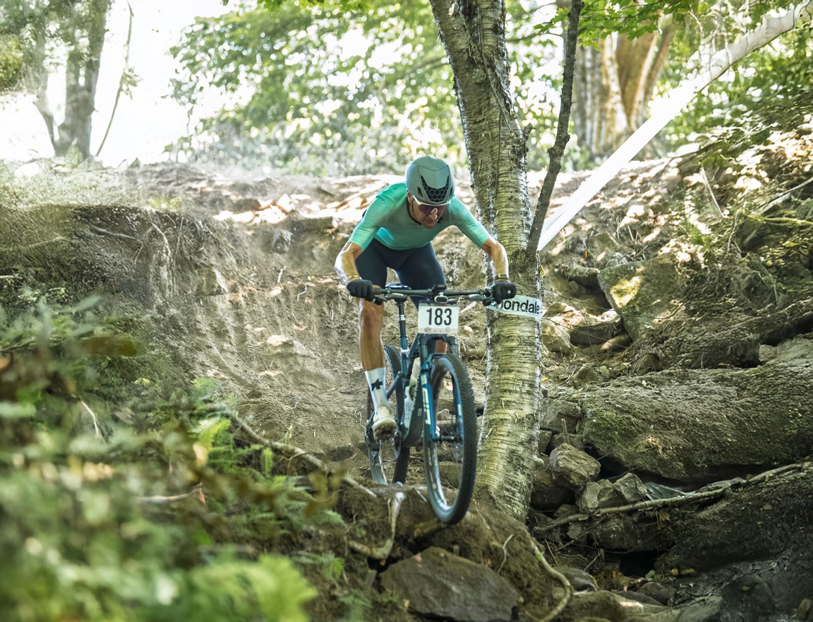 Cyclist Ted King navigating rocky, wooded terrain. Number 163 visible. Wearing green jersey.
