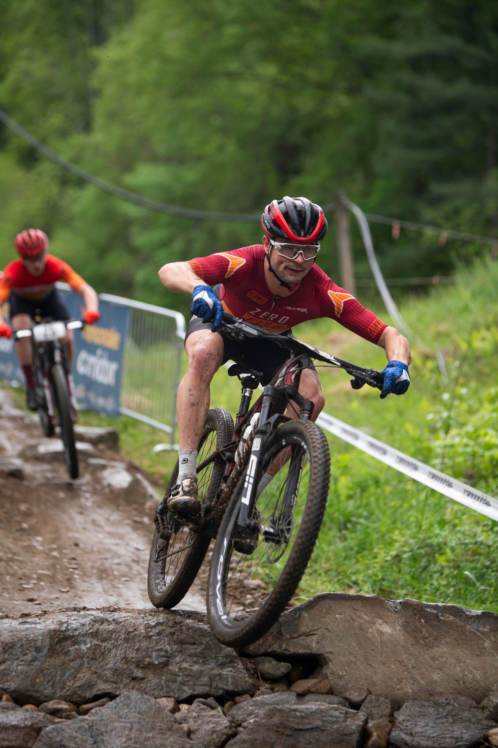 Mountain biker navigates rocky terrain during a race. Red jersey, focused expression, wooded setting.