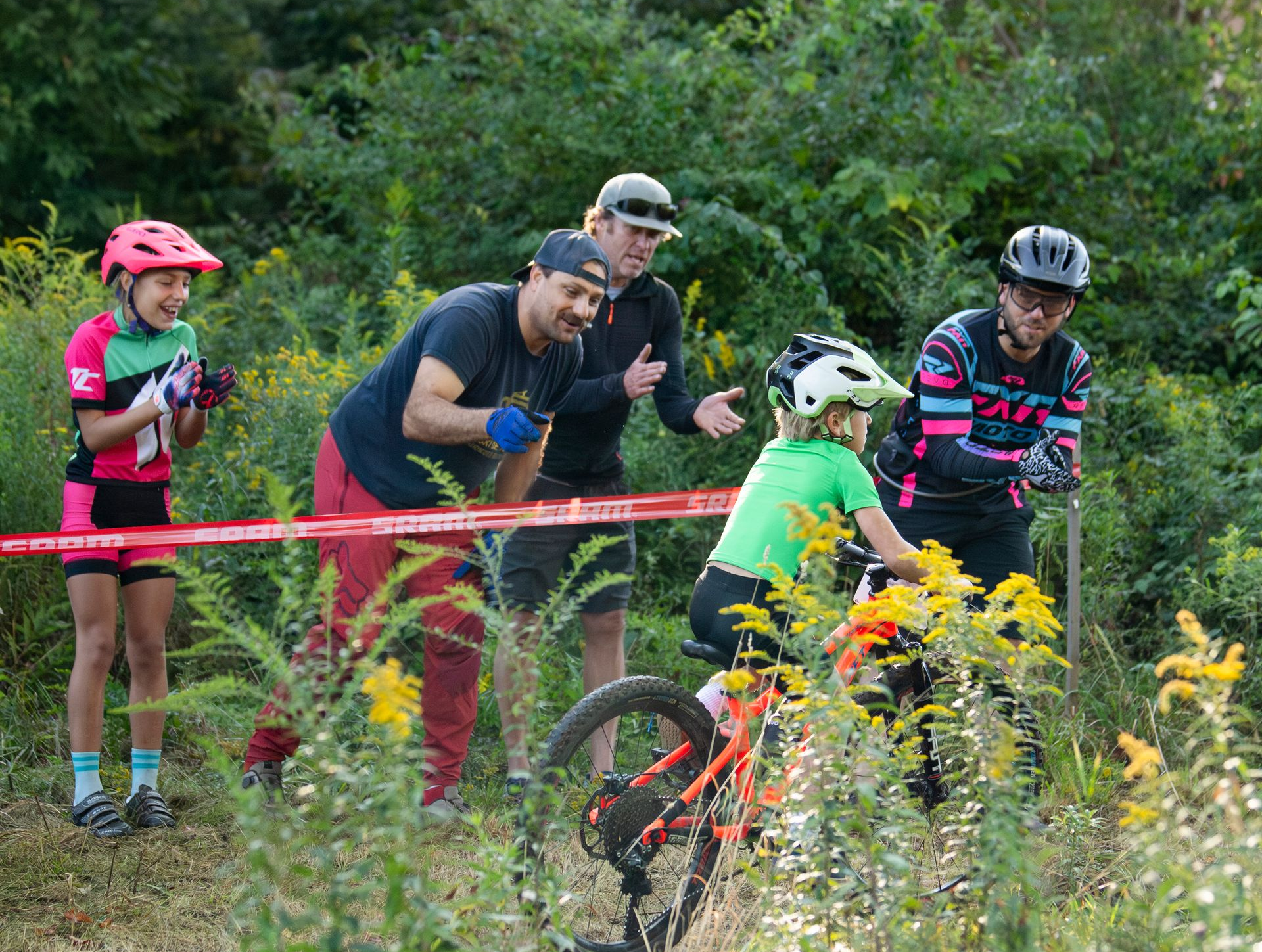 People watching a child on a mountain bike race through a grassy area. Cheering, red ribbon barrier.