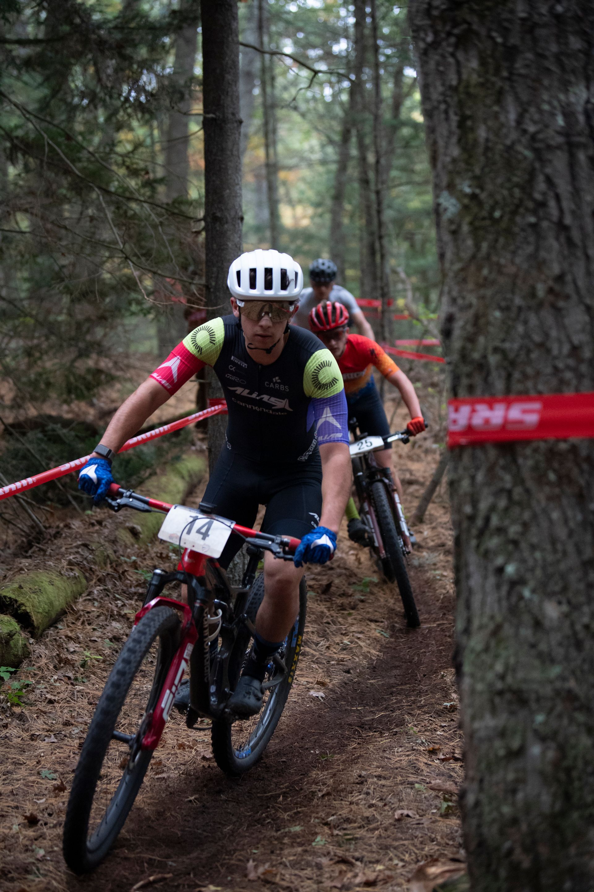 Mountain bikers racing on a dirt trail in a forest, trees and red course tape visible.