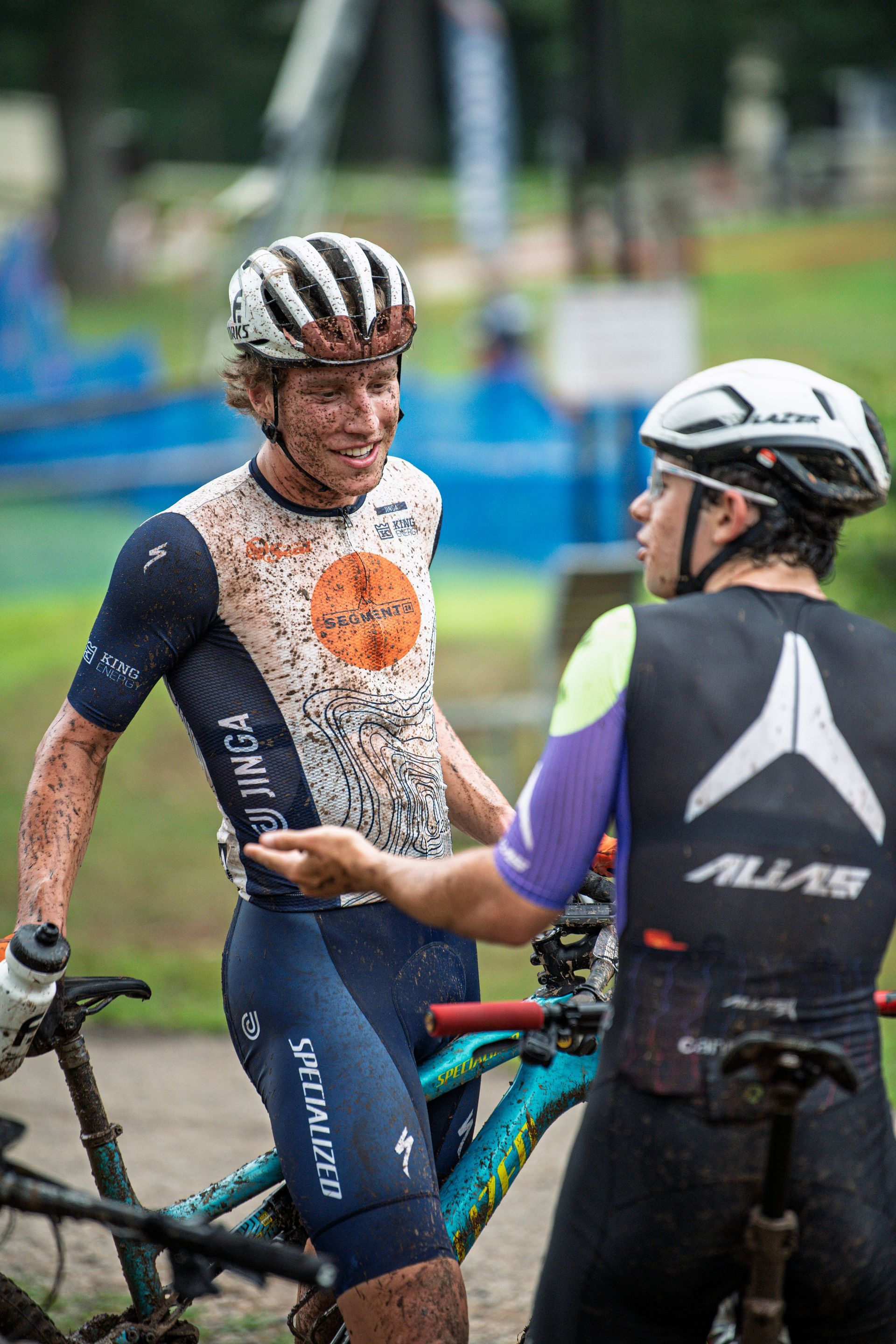 Cyclist covered in mud smiles at another person; mountain bike visible; outdoor setting.