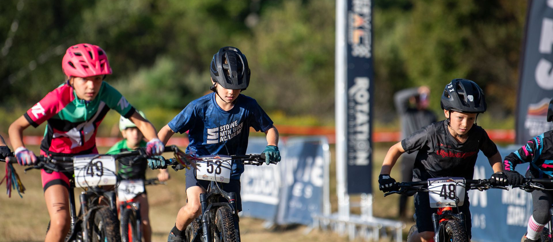 Kids on bikes racing on a dirt track, wearing helmets and athletic gear.