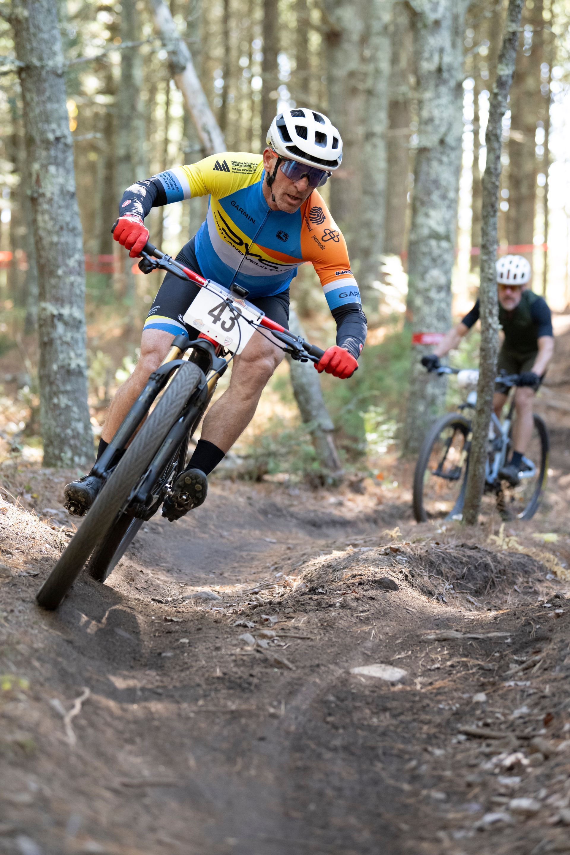 Cyclist in colorful jersey leans into a turn on a dirt path, number 43 visible. Another cyclist follows. Forest setting.