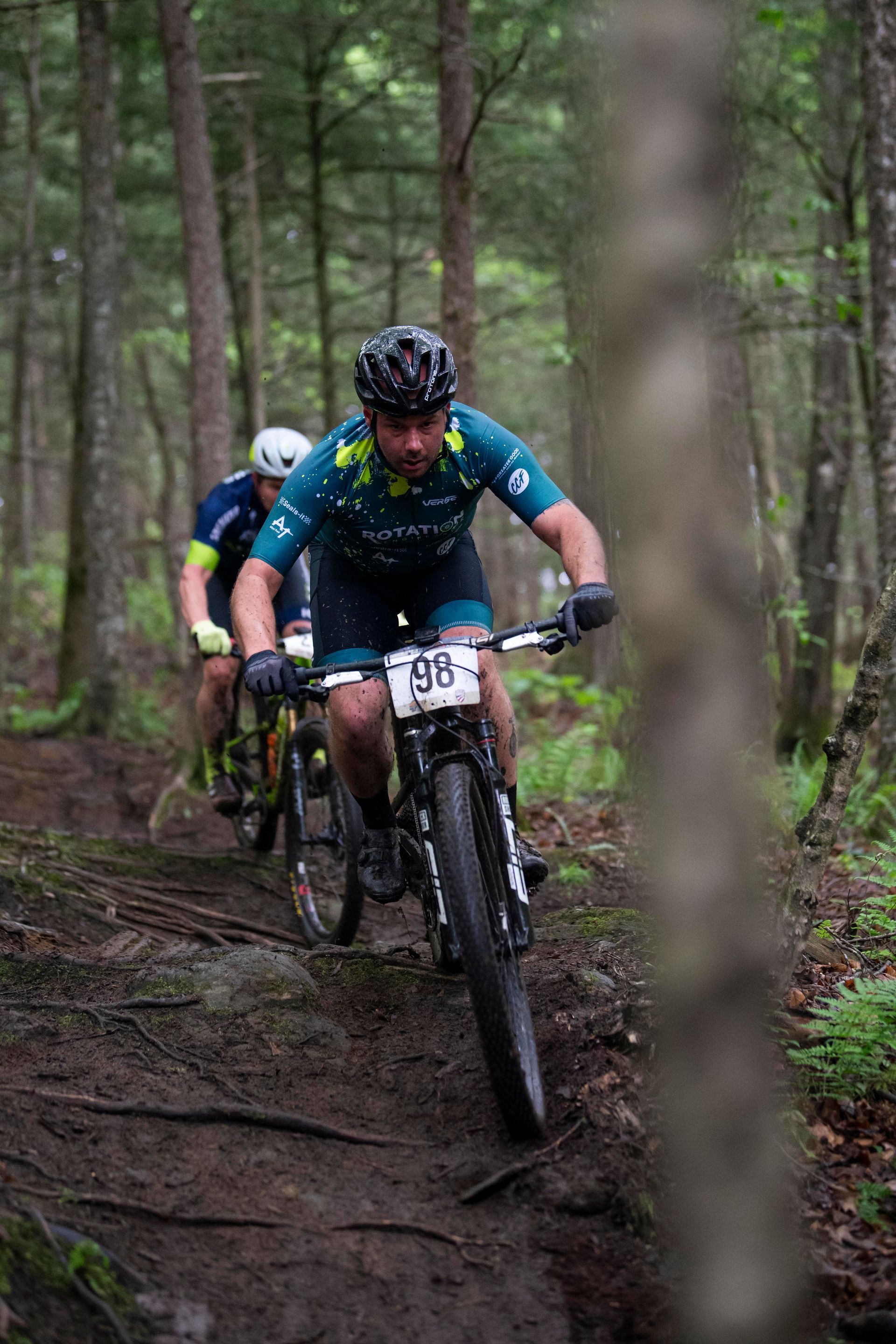 Mountain bikers racing on a muddy trail in a forest, the lead rider wearing a teal jersey, number 58.