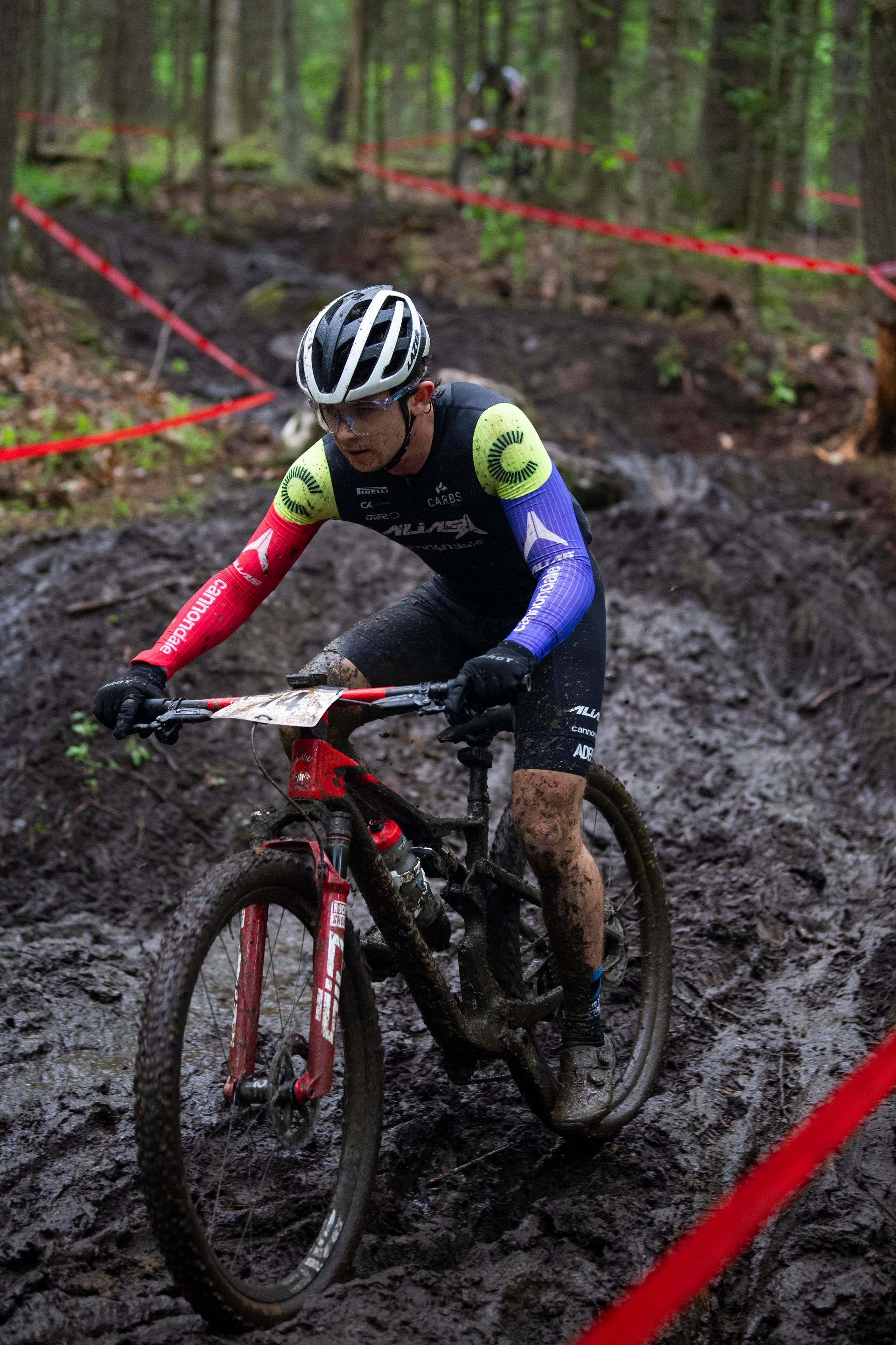 Mountain biker in black and colorful jersey pedals through muddy terrain in a forest.