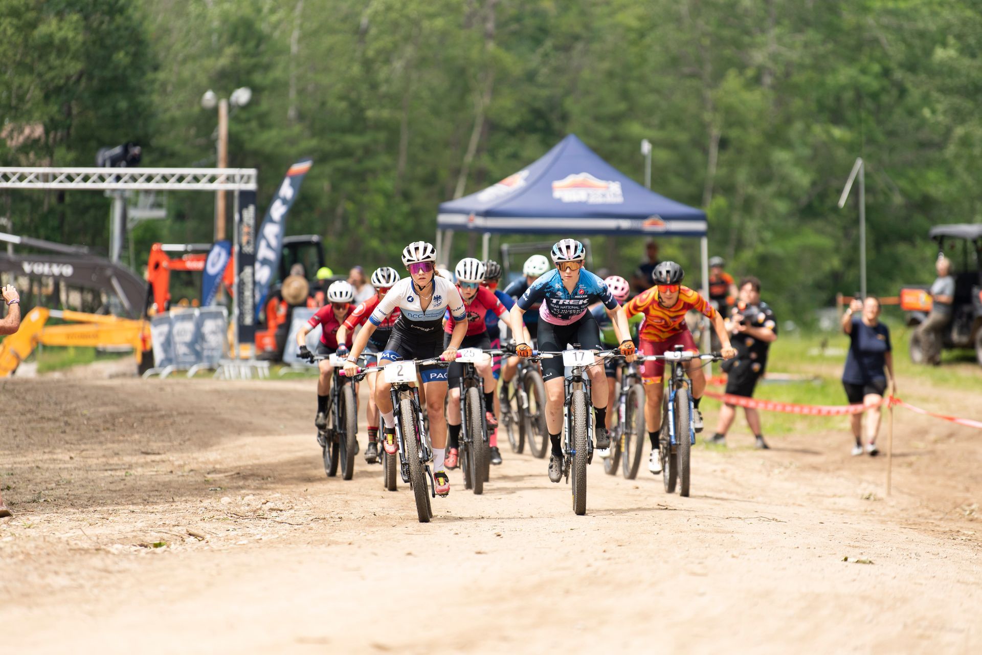 Mountain bike race on a dirt course; riders in jerseys race toward the finish line, spectators nearby.