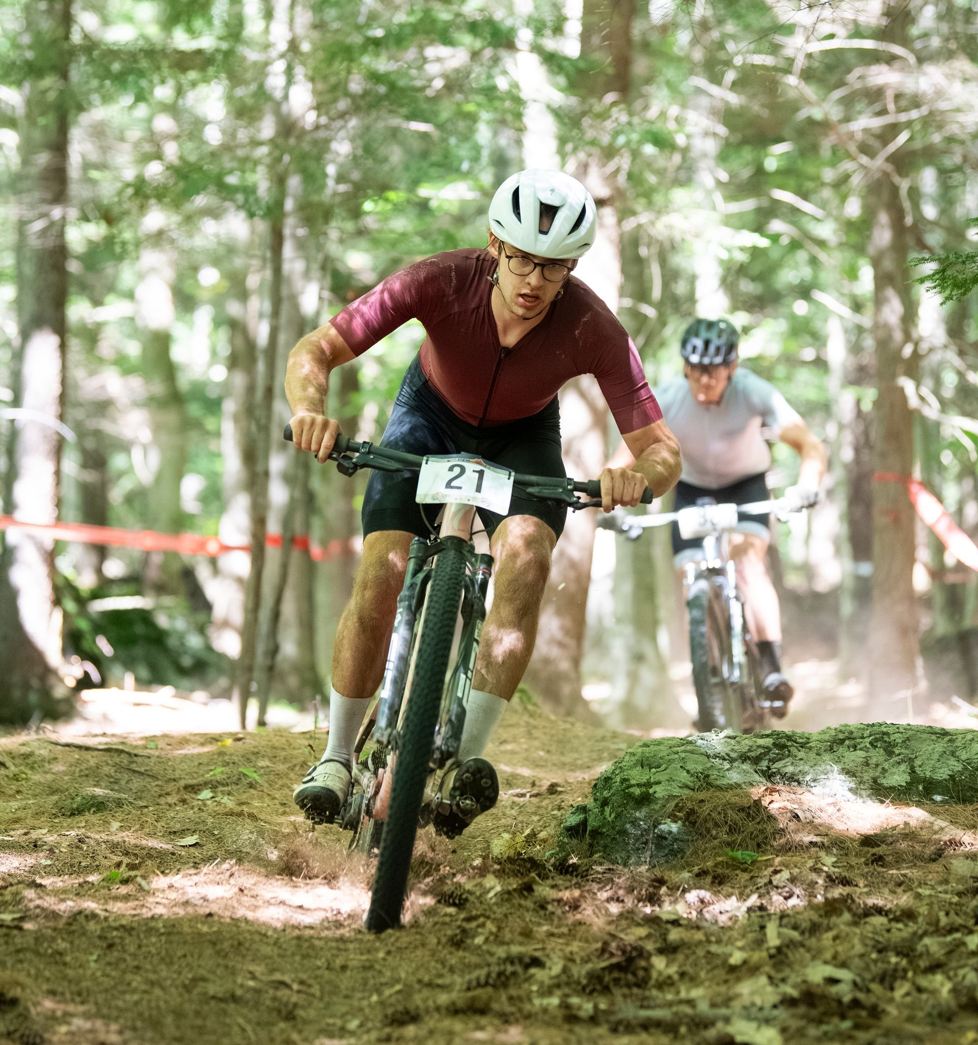 Mountain bikers competing on a wooded trail; front rider in maroon, number 21, and white helmet.