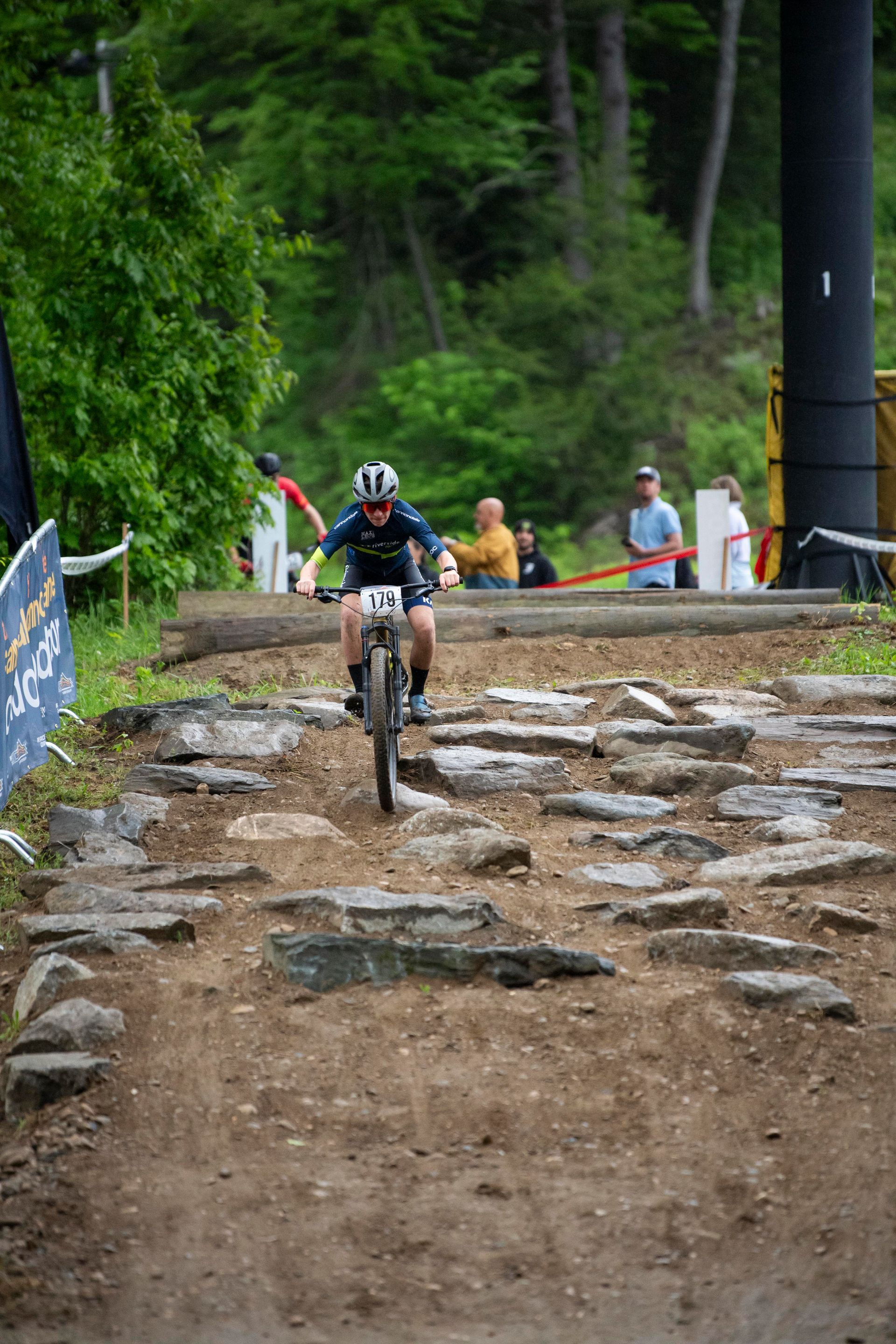 Mountain biker navigates a rocky trail, focusing ahead. Forest setting with spectators visible.