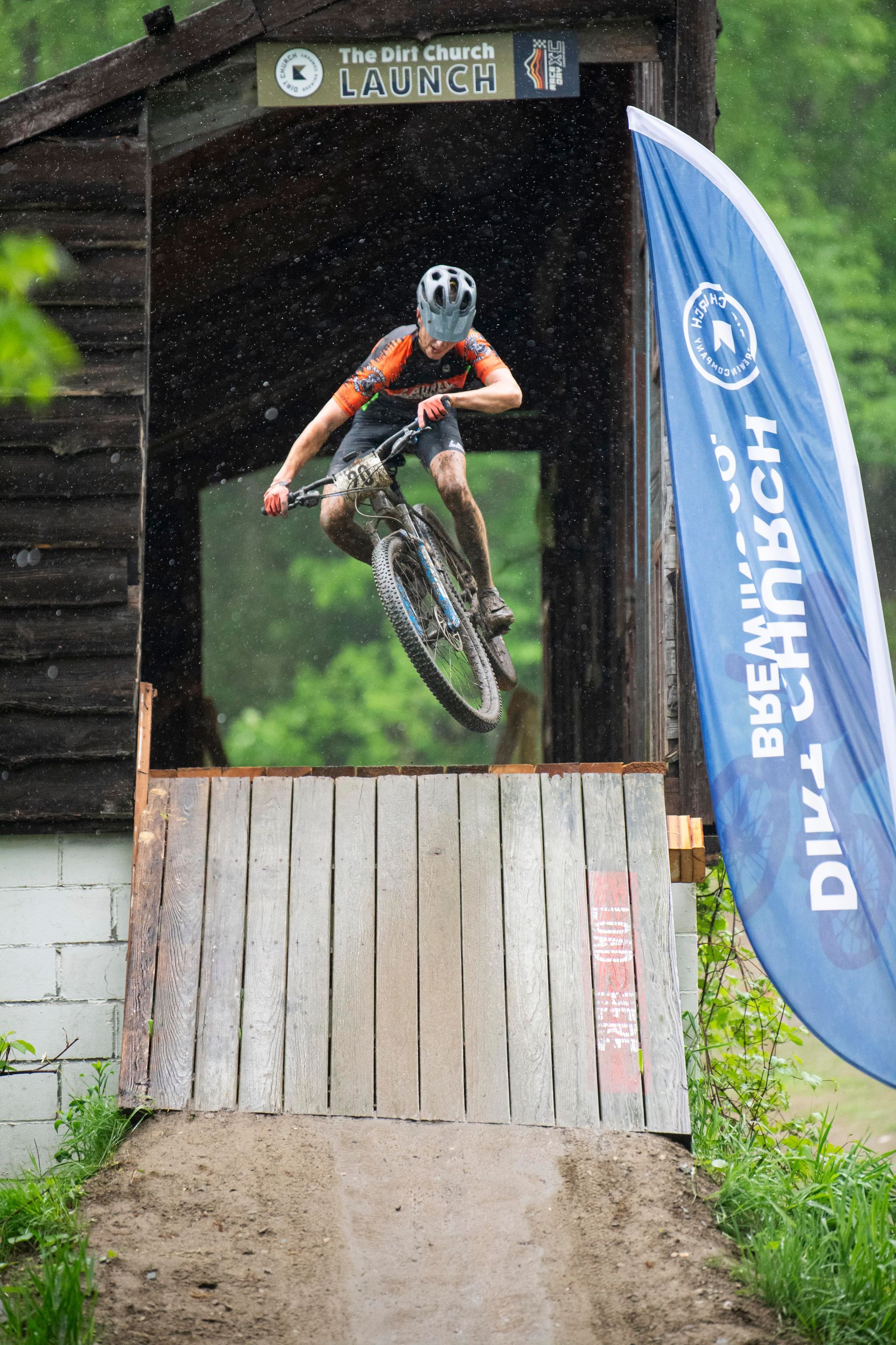 Mountain biker jumps over a wooden ramp in front of a covered bridge in the rain.
