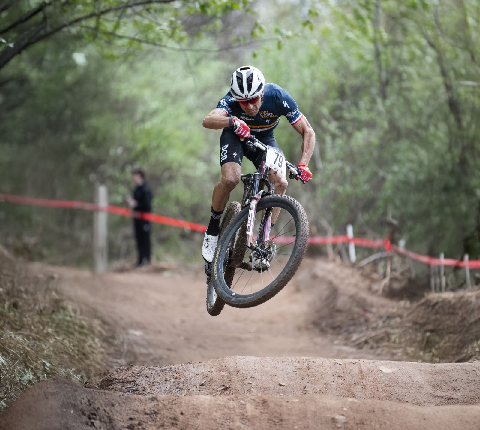 Mountain biker in mid-air over dirt jump on wooded trail, wearing a helmet and racing kit.