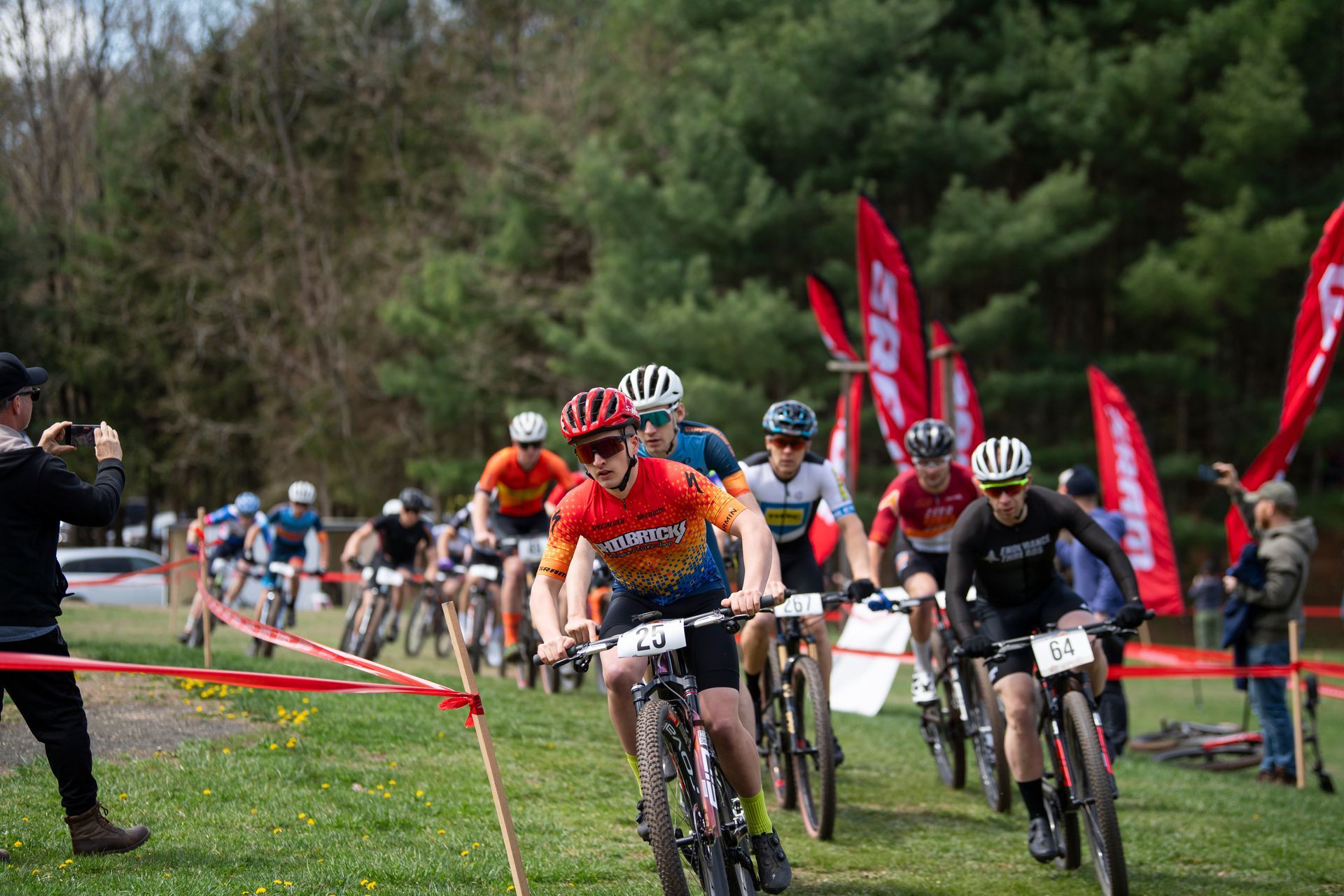 Mountain bikers racing on a grassy course, red flags in background.