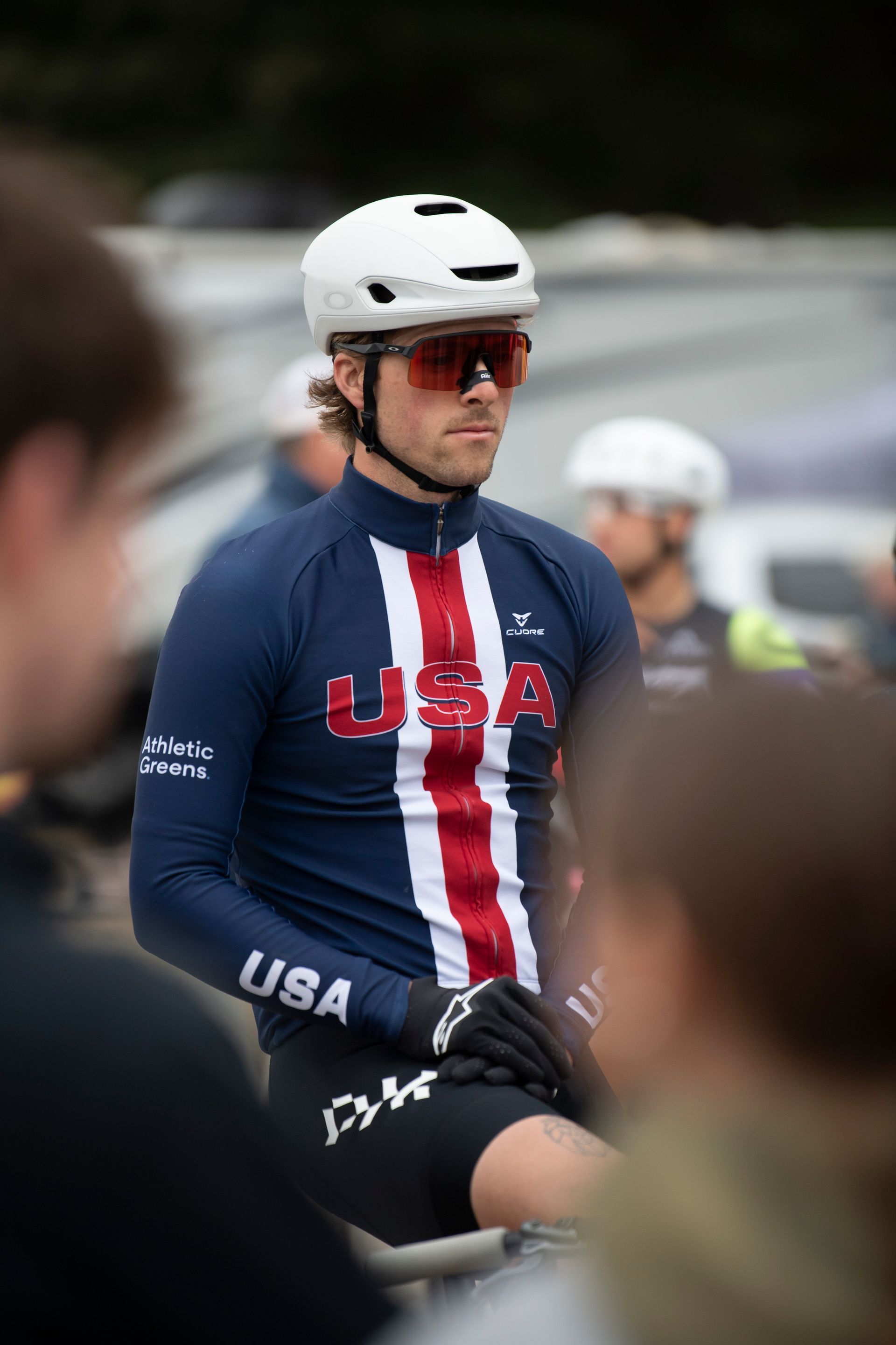 Cyclist in USA jersey, white helmet, and sunglasses, looking intently, outdoors.