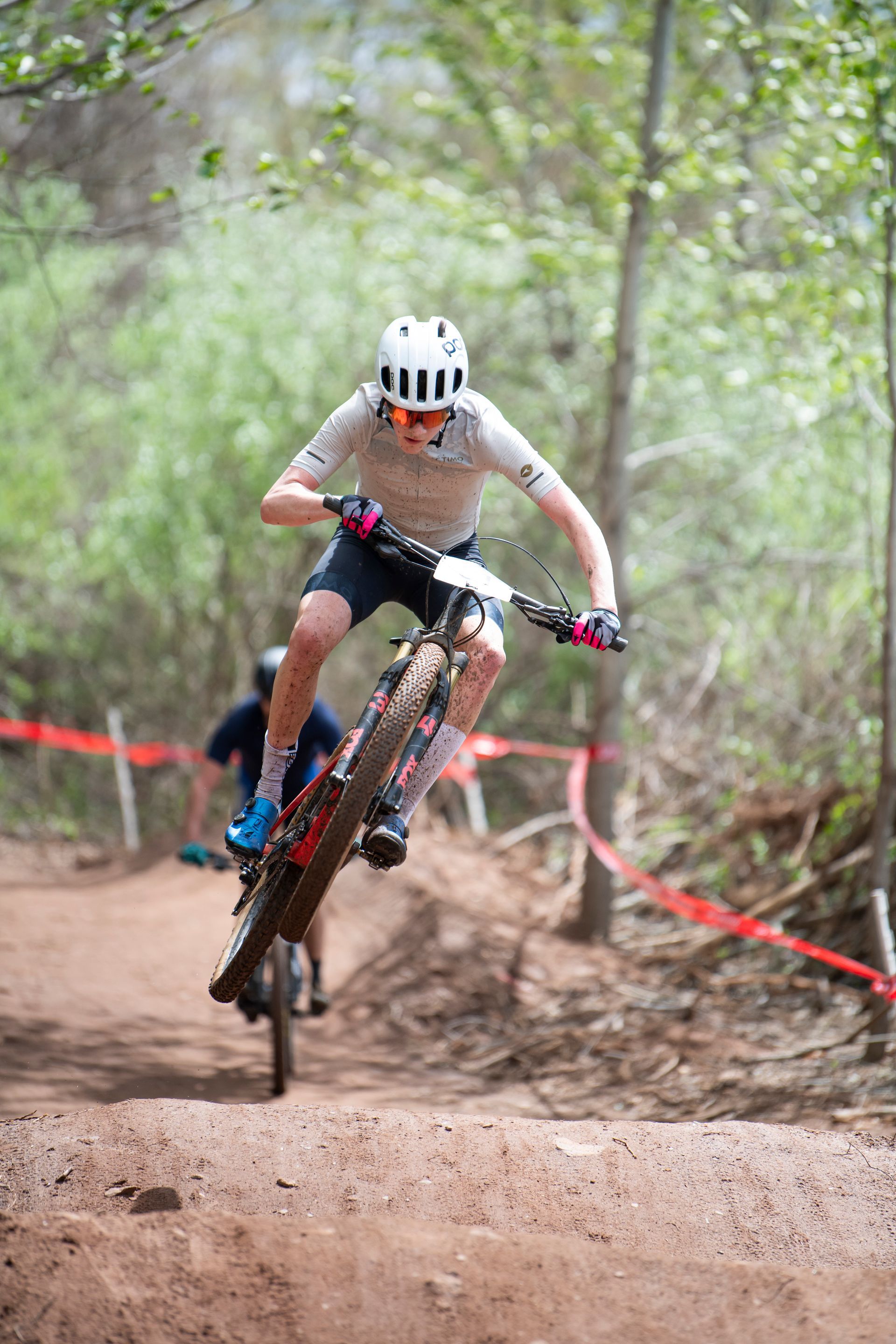Mountain biker in white helmet and jersey jumps a dirt mound on a trail; another rider follows behind.