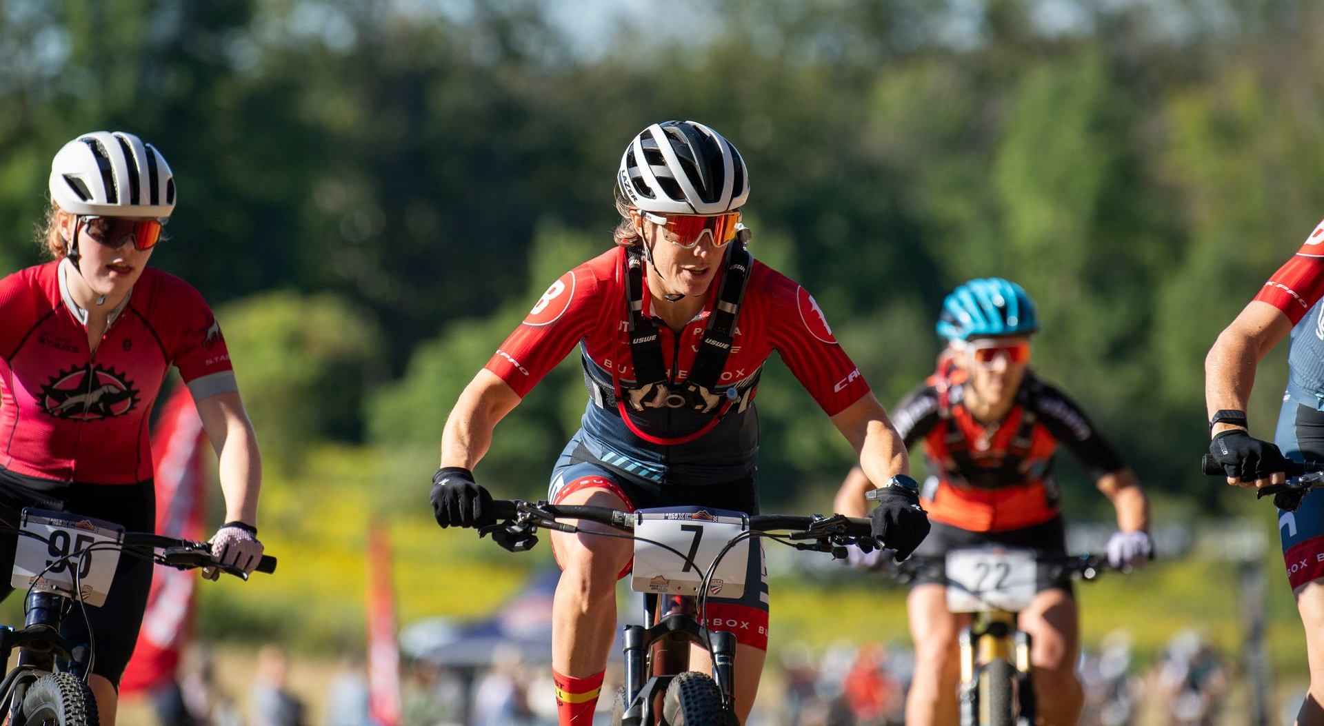 Mountain bikers racing on a dirt path in bright sunshine; riders in helmets and athletic wear.