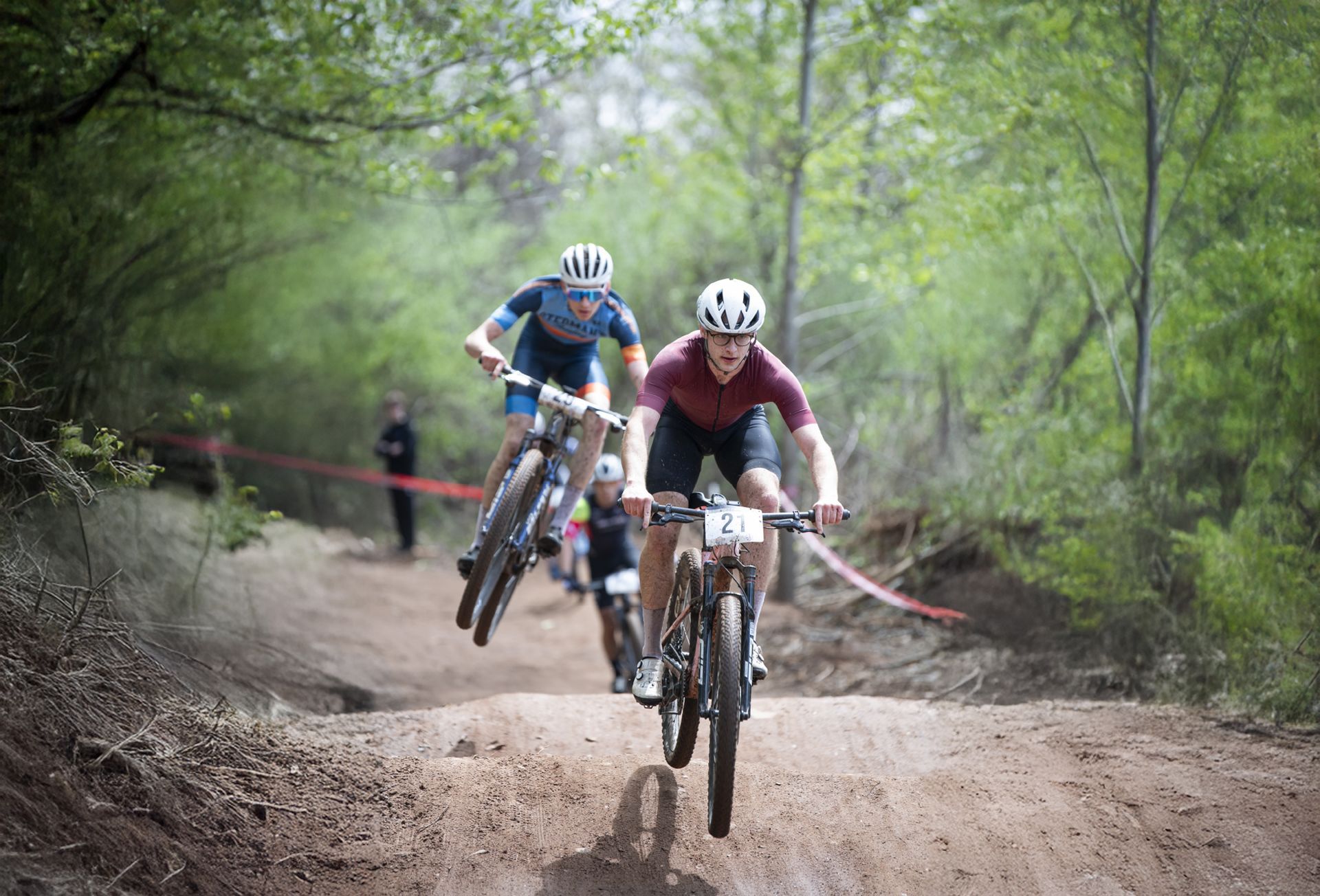 Mountain bikers racing on dirt trail, jumping over a small hill.