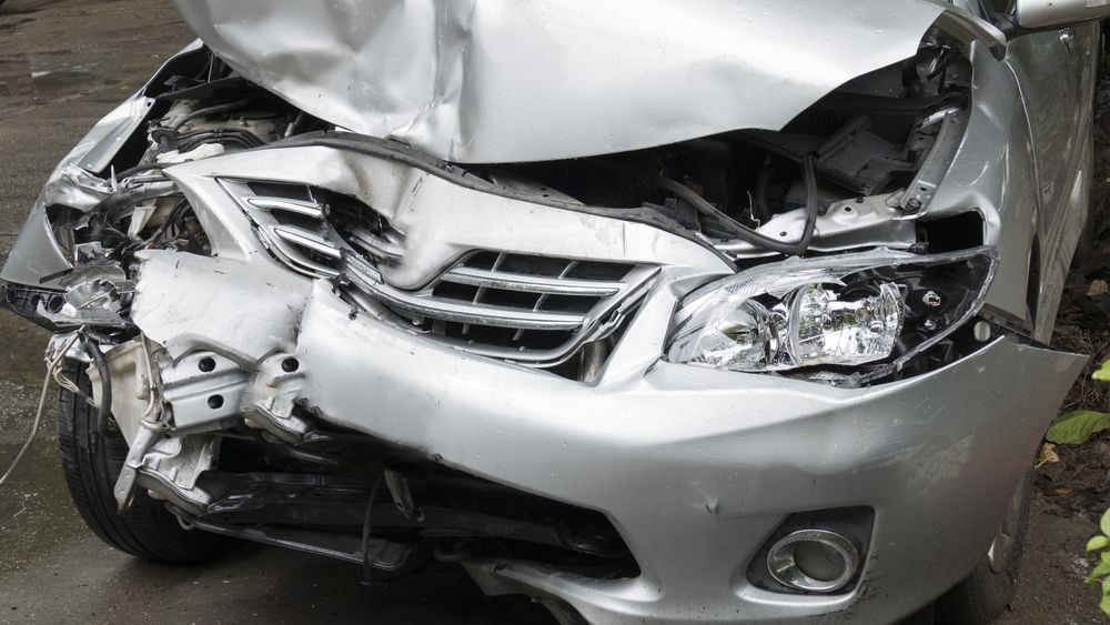 Damaged silver car front end; crumpled hood, headlights, and bumper after a collision.