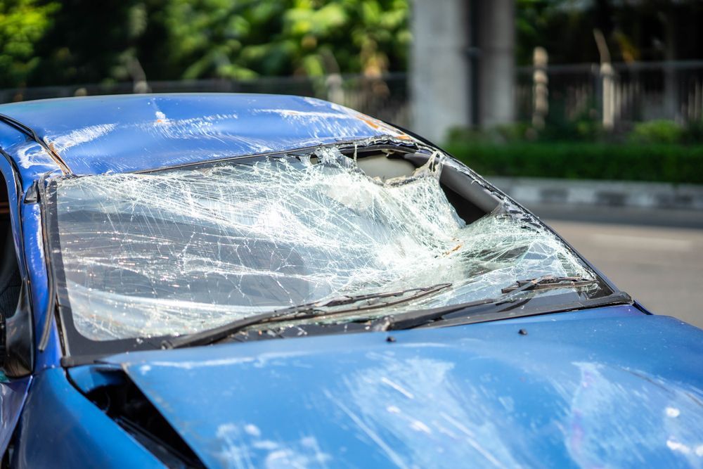 Blue car with a shattered windshield, front end damaged, in a street setting.
