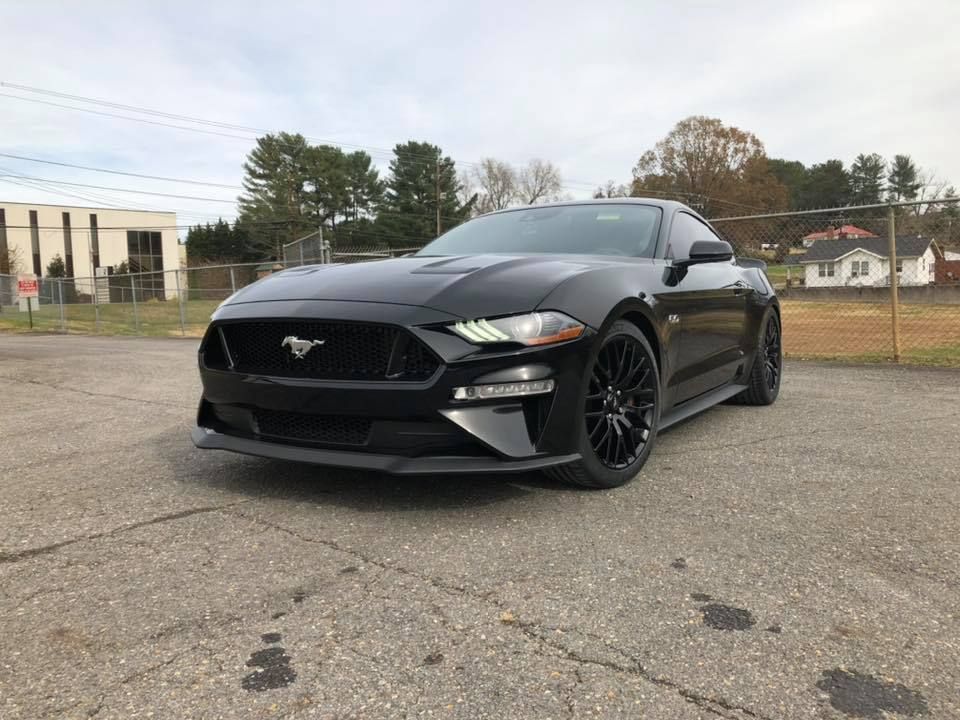Black Ford Mustang parked on an asphalt lot. Trees and a building are in the background.