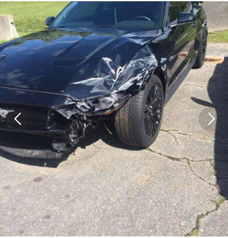 Damaged black Ford Mustang car with front-end collision on paved road.