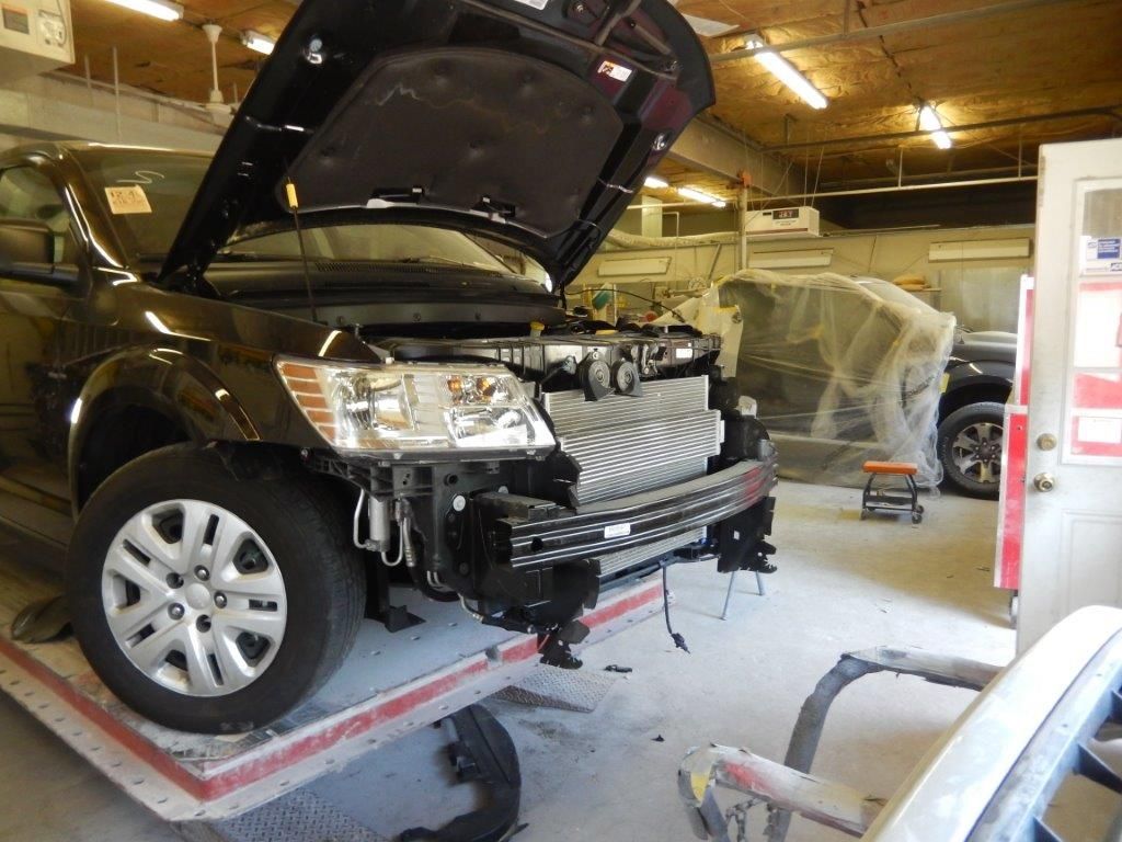 Black car in auto repair shop with hood open, front bumper removed.