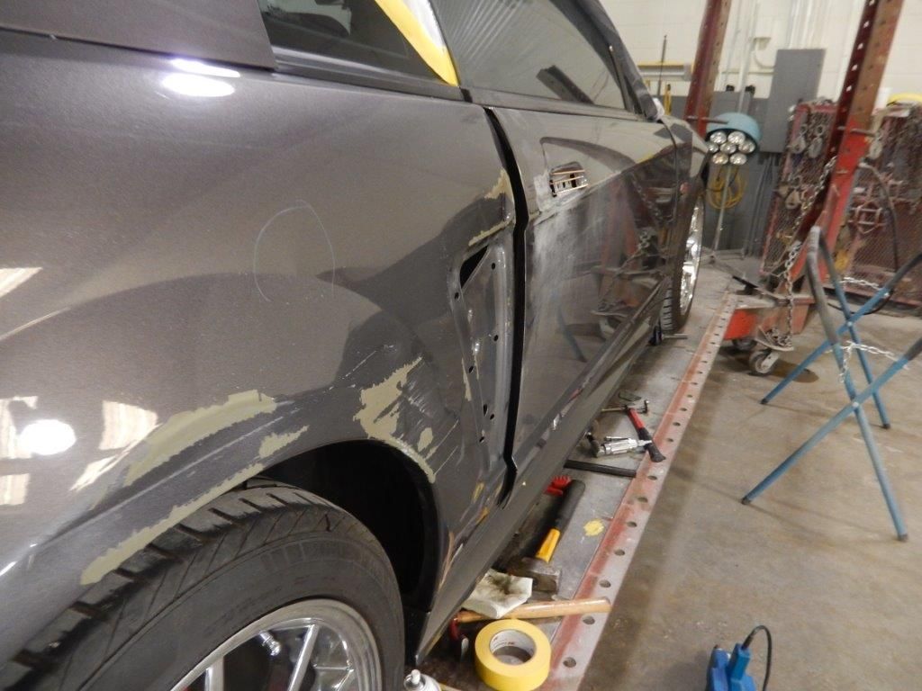 Car with damaged door and quarter panel in a repair shop, partially stripped of paint.