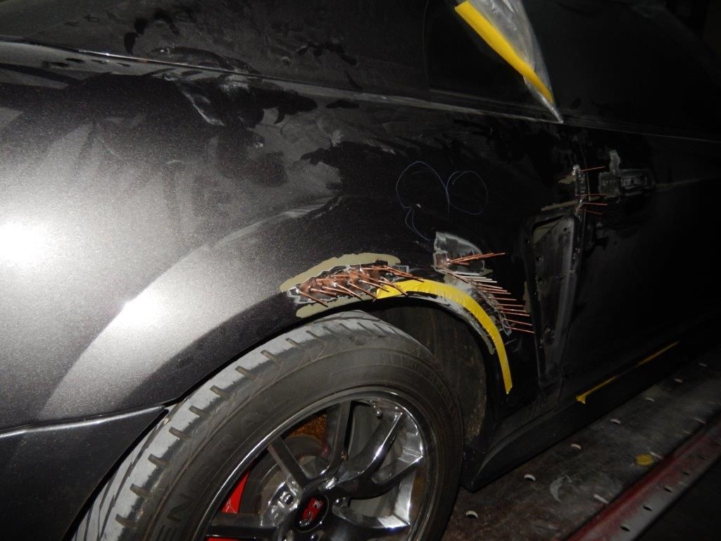 Damaged black car's rear quarter panel with exposed metal and yellow tape, next to a tire.