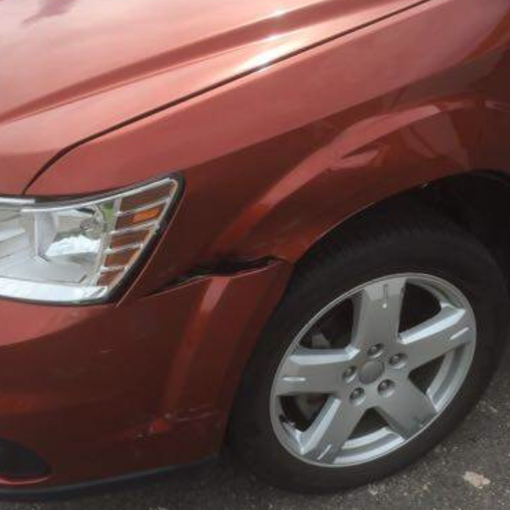 Damaged front right of a red car, dented fender, cracked paint, headlight and wheel visible.