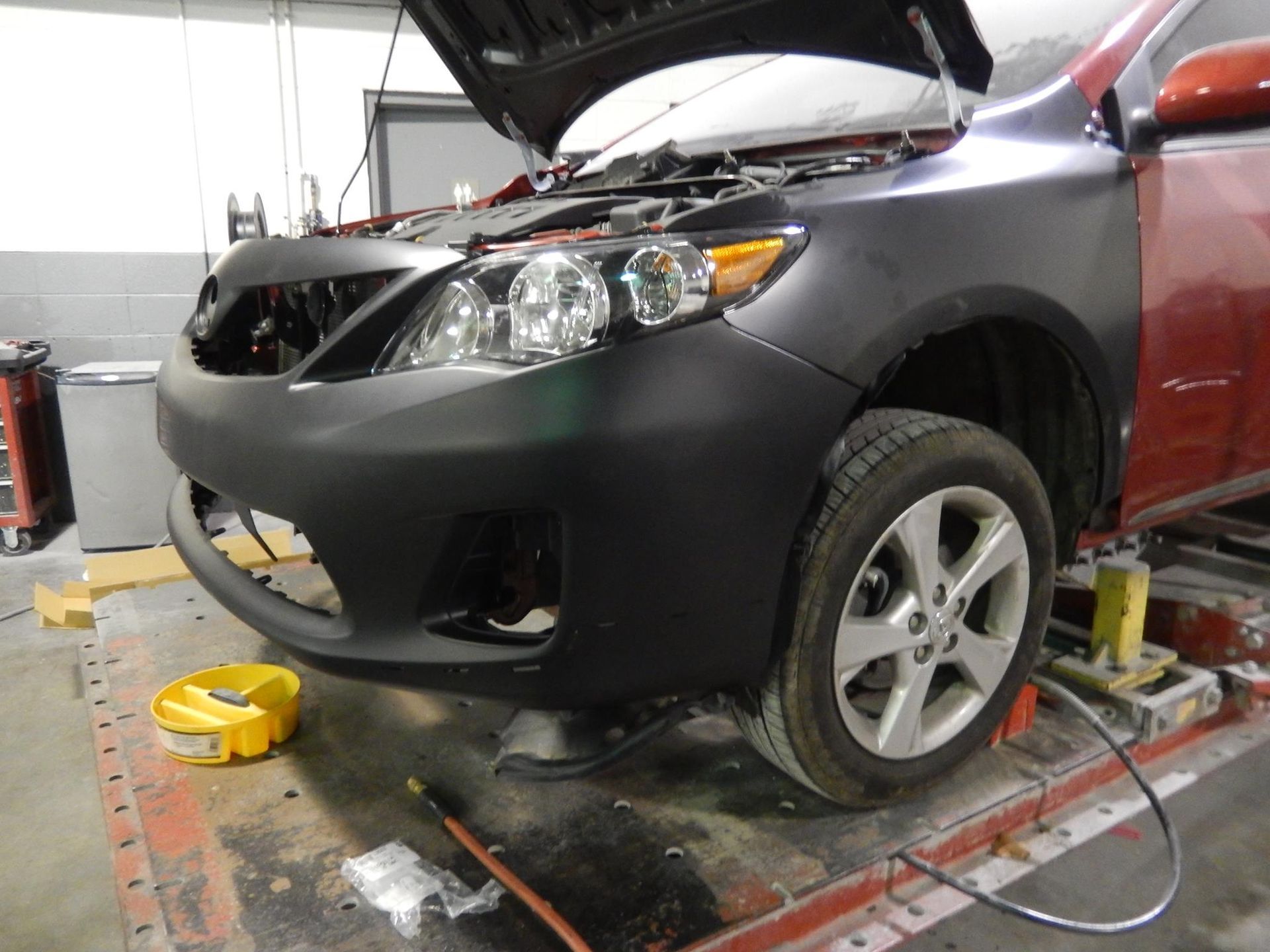 Car with hood open, front end repaired with new black bumper and fender, in a shop.