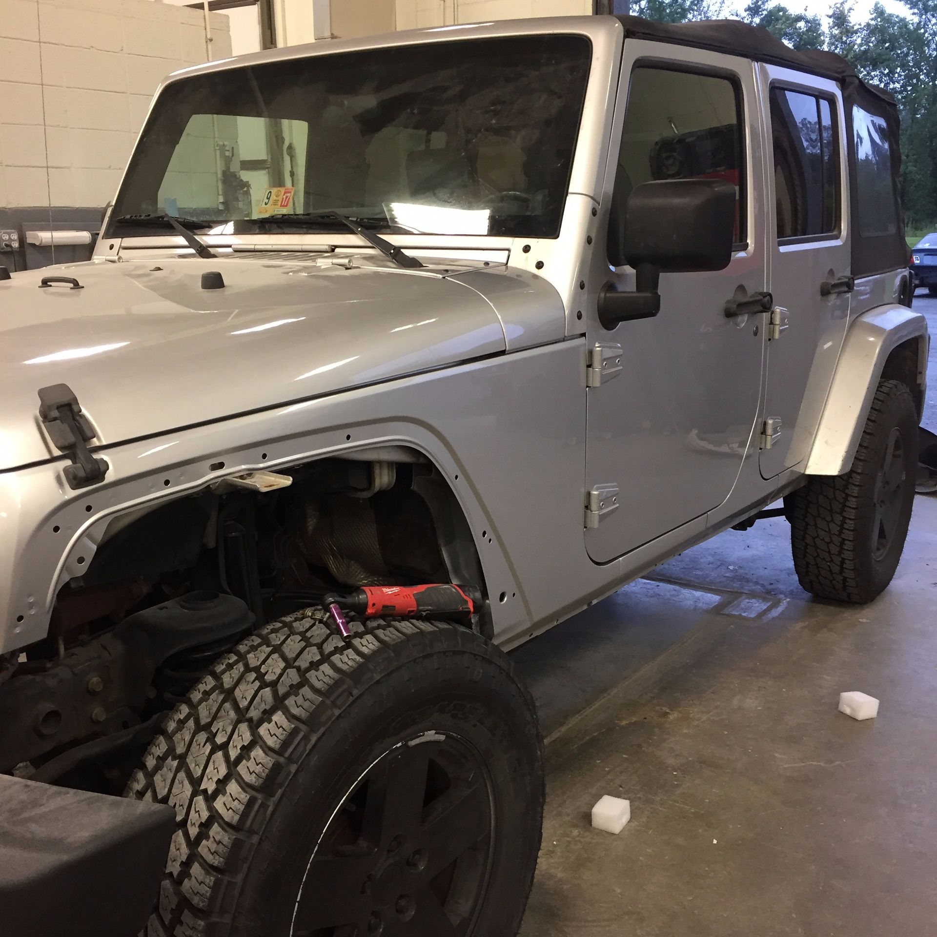 Silver Jeep Wrangler with front fender removed in a garage. Black tires and soft top.