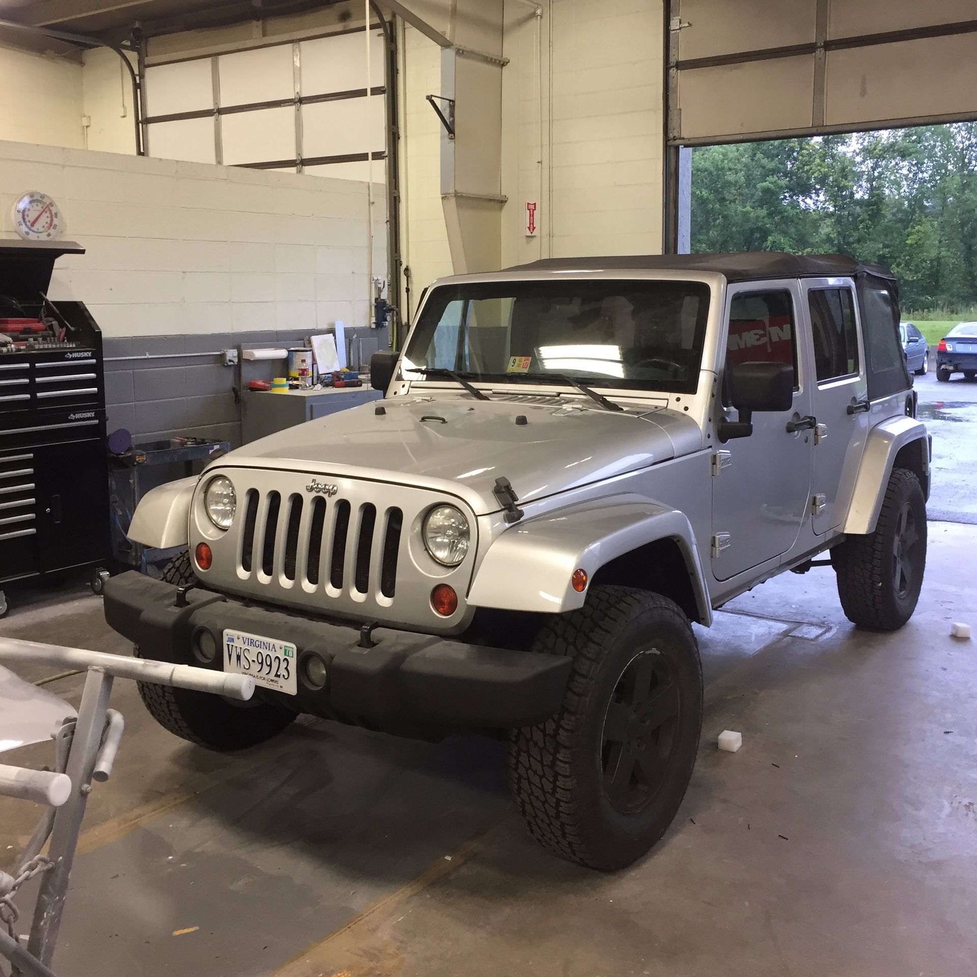 Silver Jeep Wrangler parked inside a garage, with black tires and top.