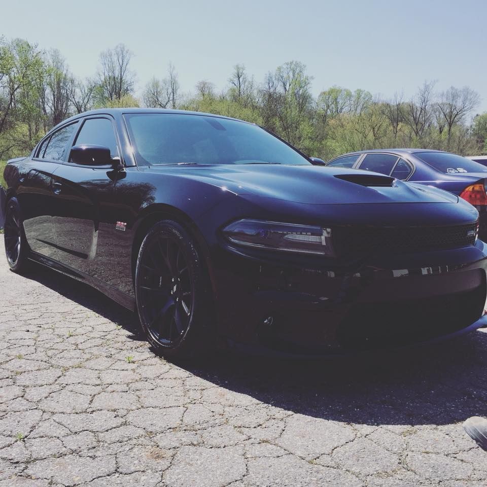Black Dodge Charger parked on a cracked pavement on a sunny day. Hood scoop and tinted windows.