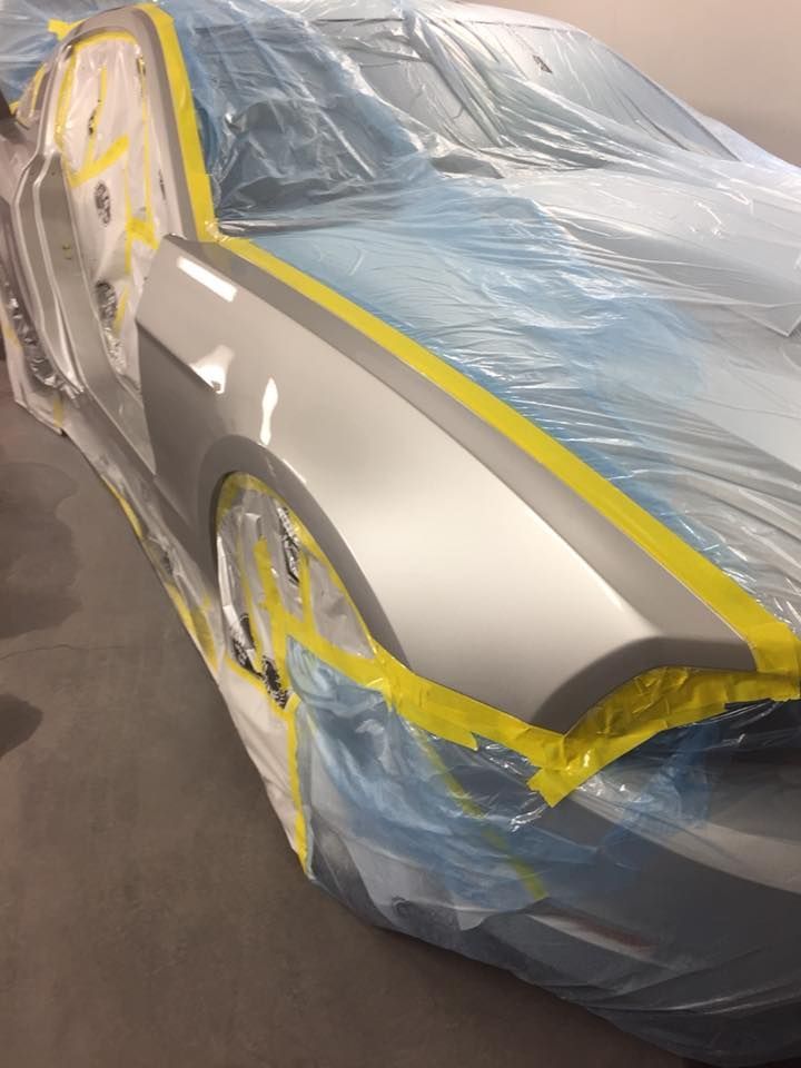 Silver car in a paint booth, taped with yellow tape, covered in blue plastic sheeting, ready for painting.