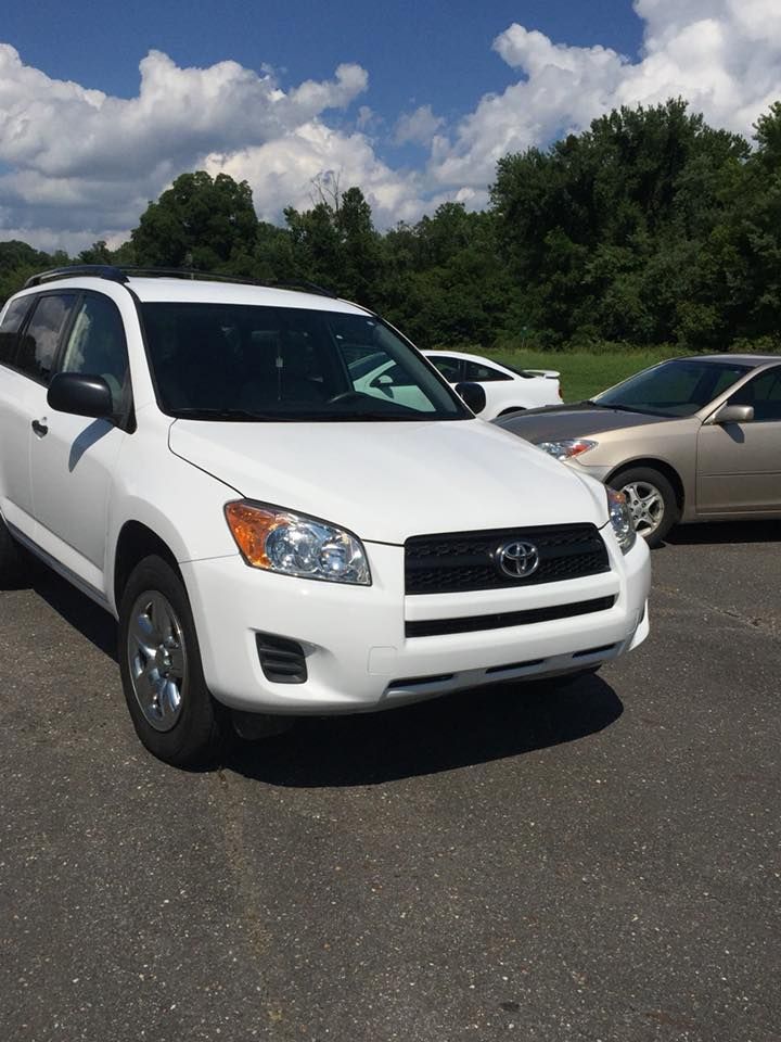 White Toyota RAV4 SUV parked on asphalt, other cars and trees in background. Bright, sunny day.